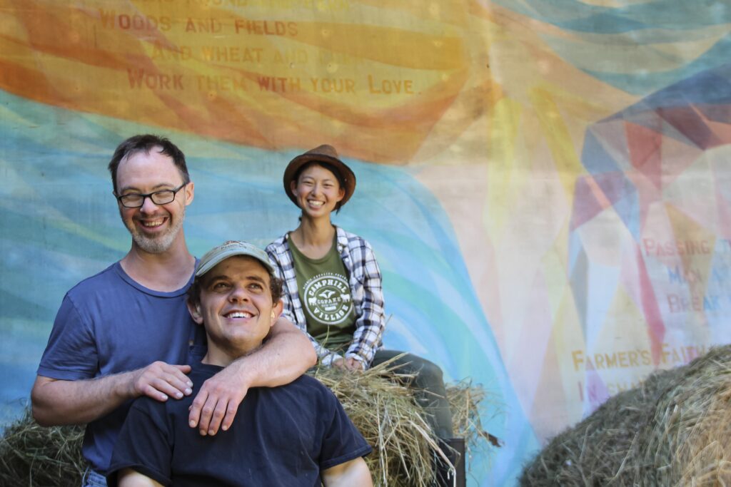 Three farmers sit together in hay bales, smiling at the camera.
