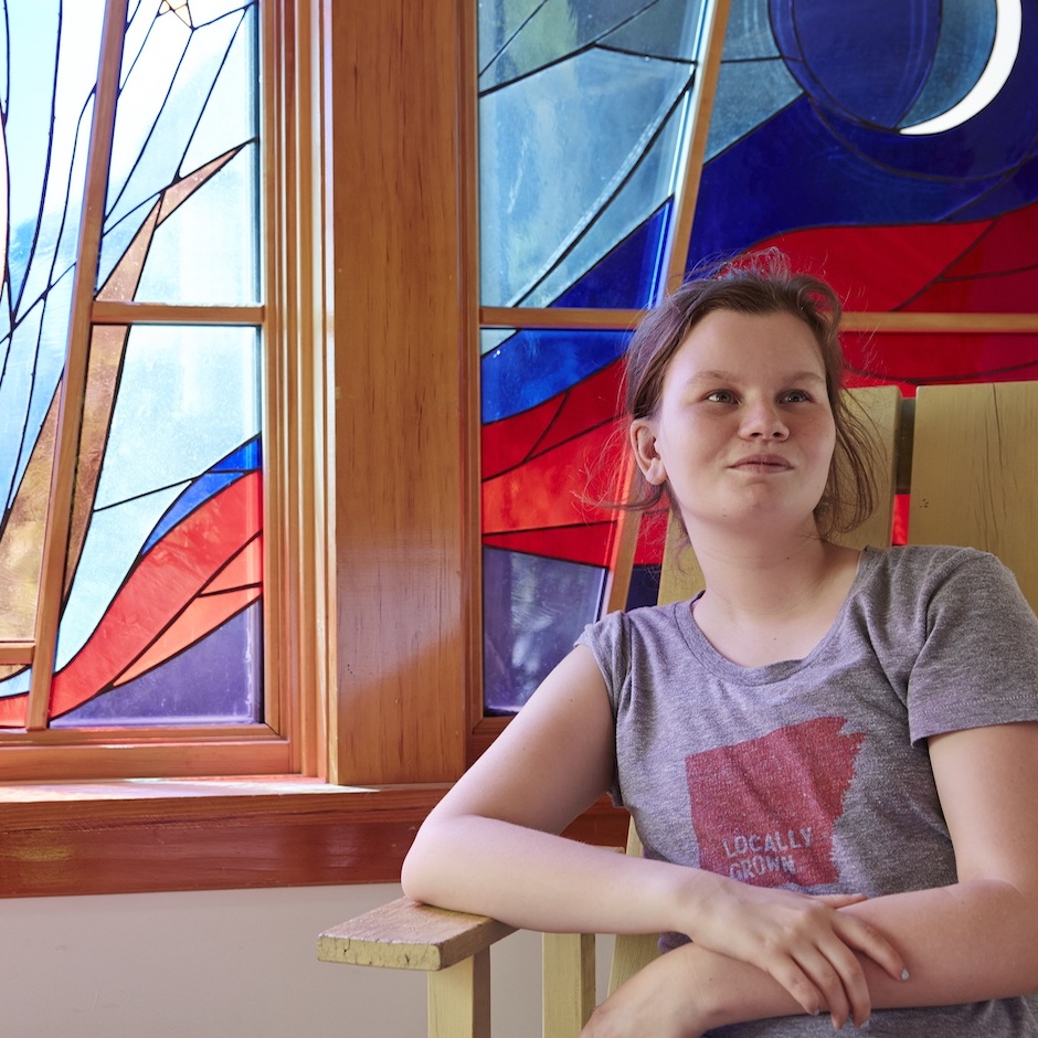 A girl sits in a chair in front of a stained-glass window, looking off to the side.