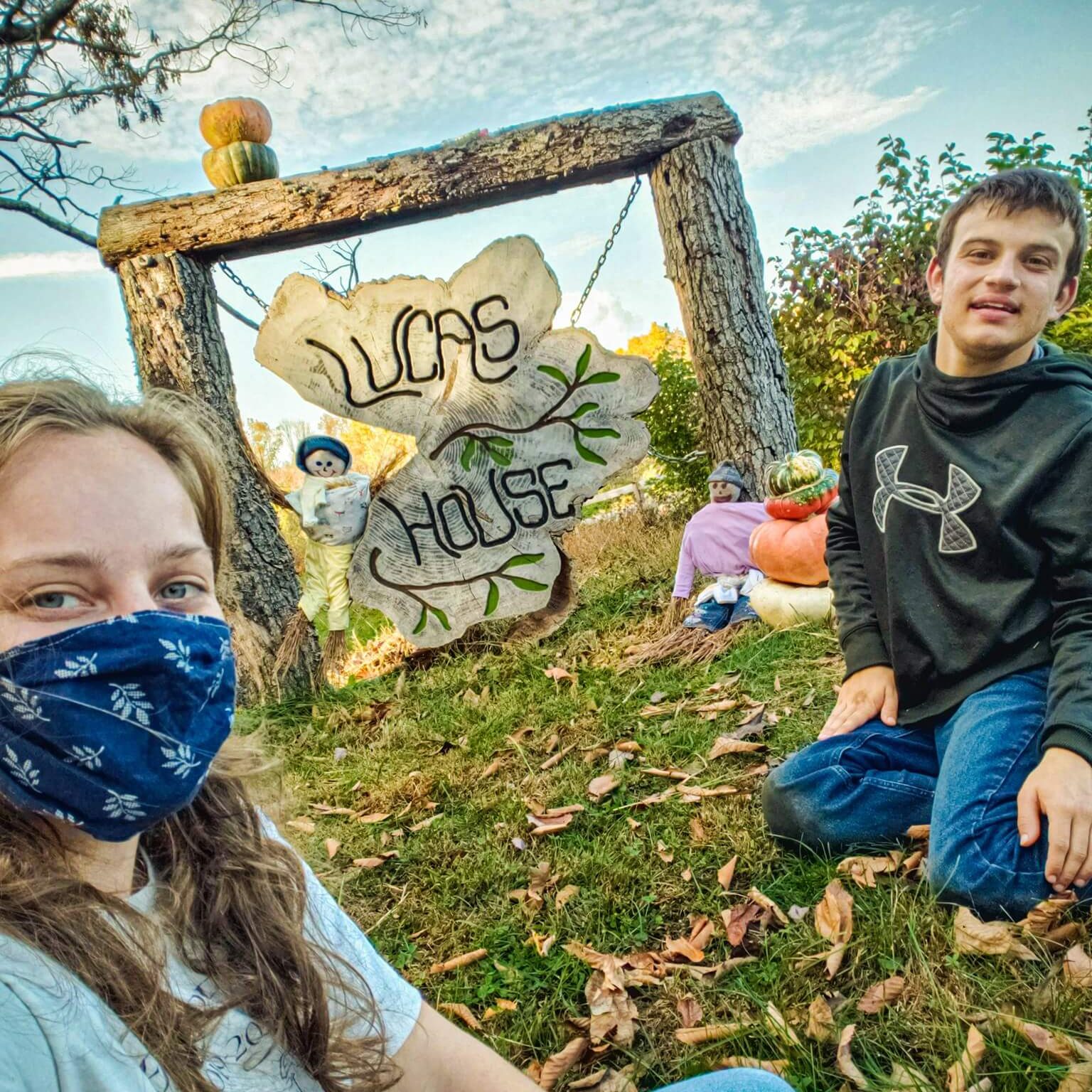 Two people posing in front of the Halloween decorations around the Lucas House sign: two handmade scarecrows and stacks of colorful pumpkins.