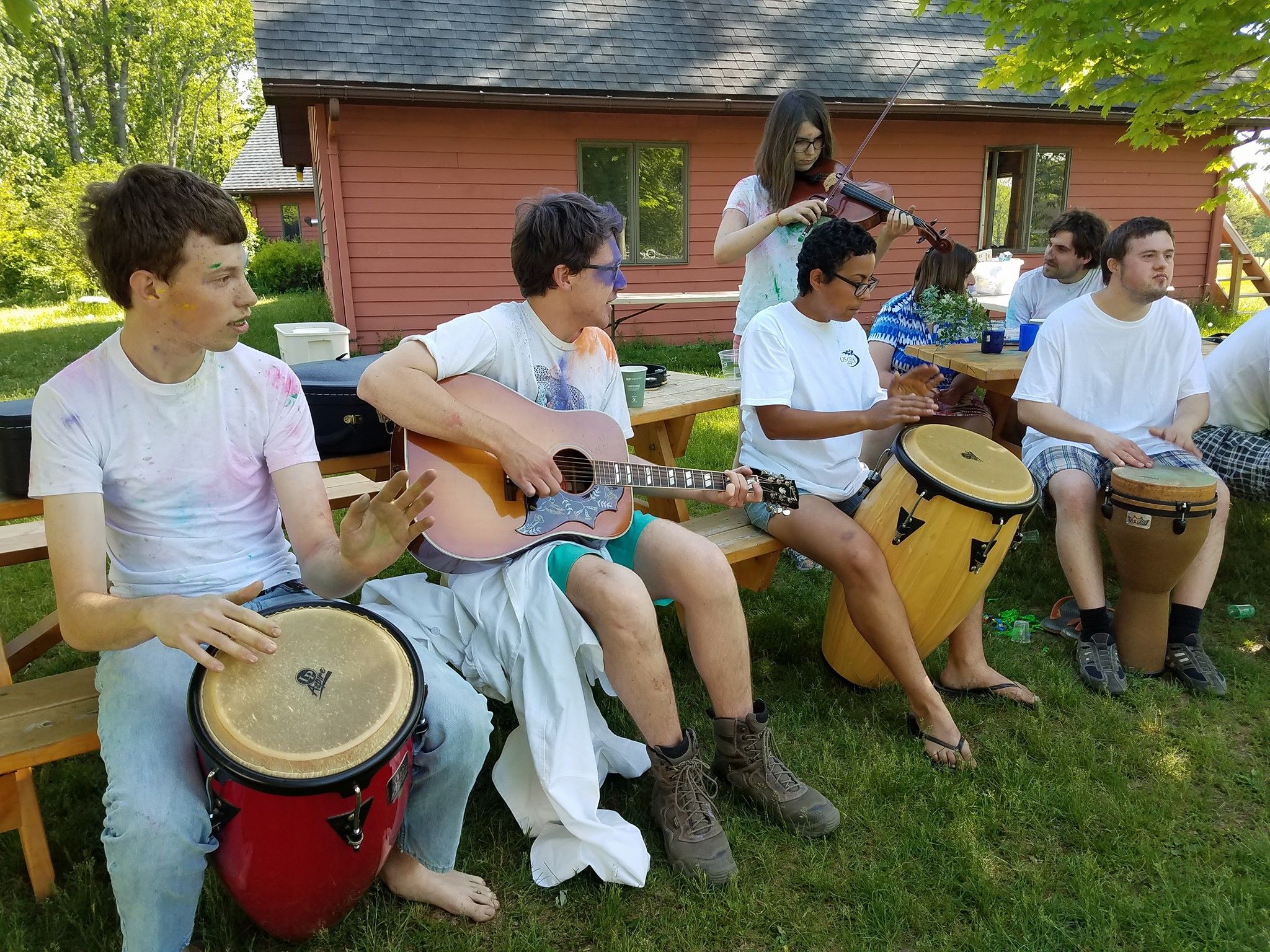 A group of drummers, a violinist, and a guitarist play together at picnic tables.