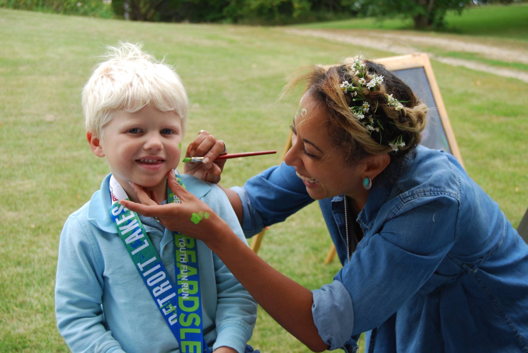 A lady with small wildflowers braided into her hair paints something lime green onto the cheek of a smiling boy.