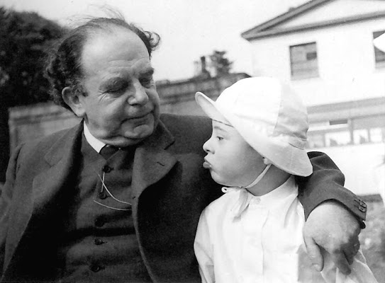 A black-and-white photo of an old man smiling down at a baby in a white hat.