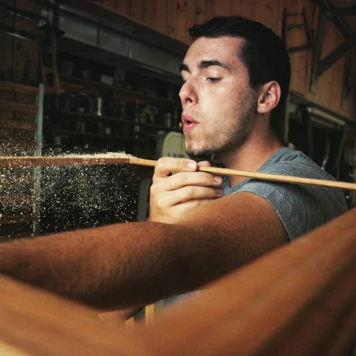 A woodworker blows sawdust off a thin piece of wood.