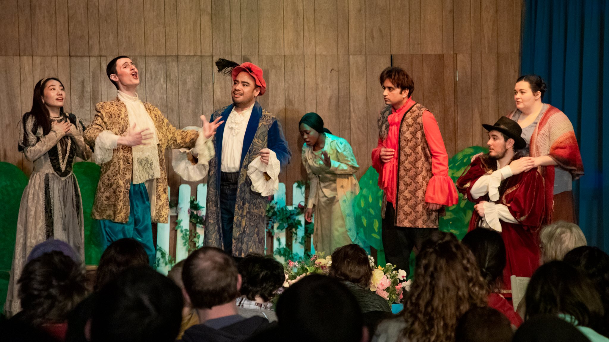 People onstage in Shakespearean costume listen in various dramatic poses as one actor delivers a speech