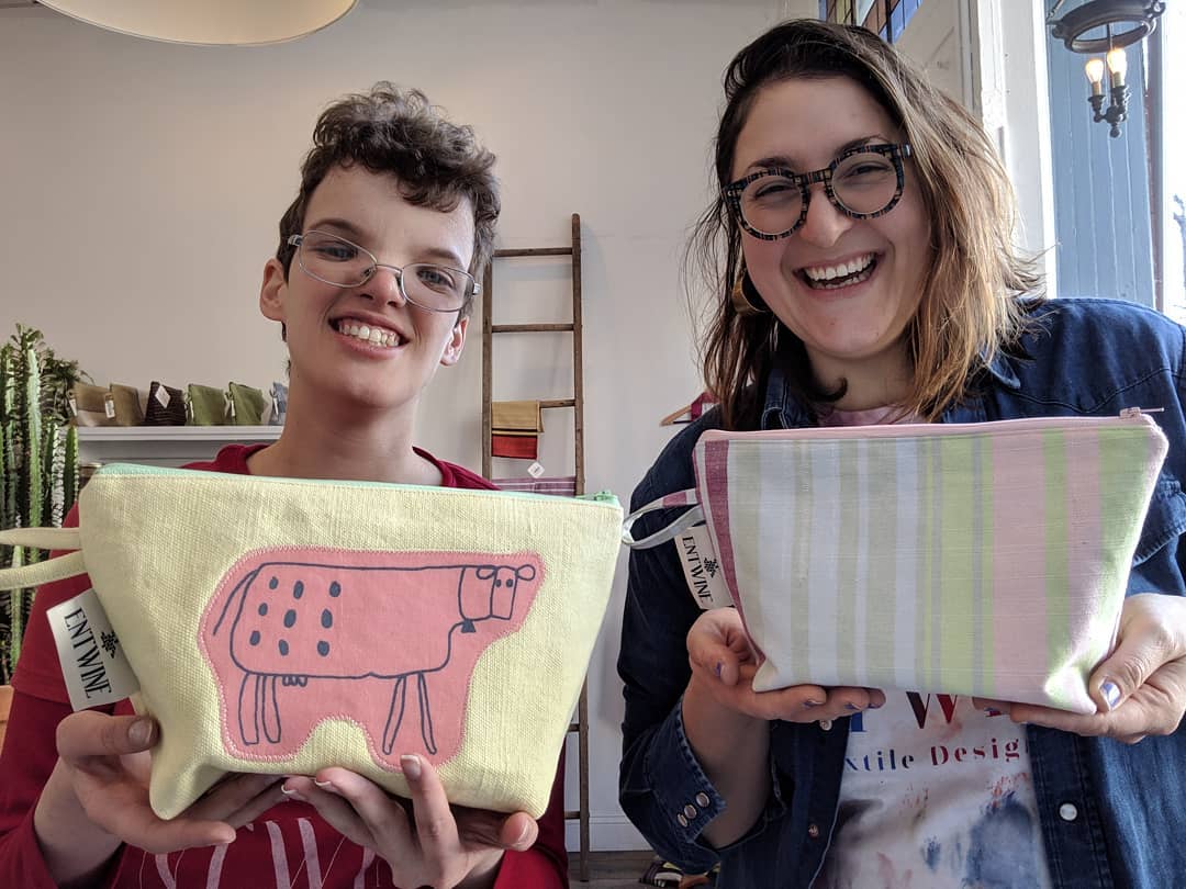 Two people hold up small zipper pouches with different designs: one with pastel stripes, the other with a sketch of a cow on pink fabric applique'd onto a solid yellow background.