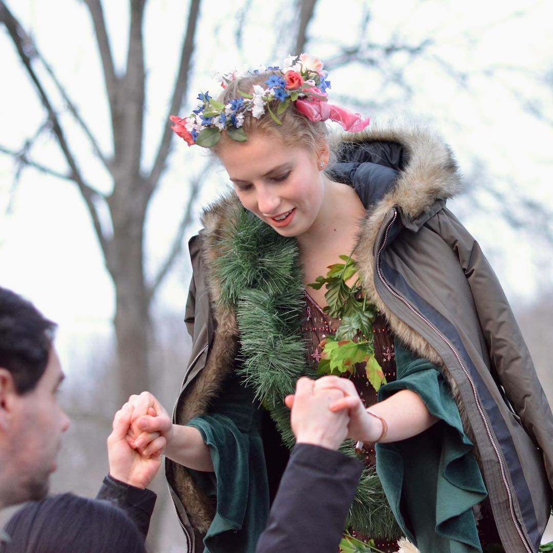 A young woman wearing a flower crown, garlands of pine needles and fake leaves, and a winter parka is helped down from a step.