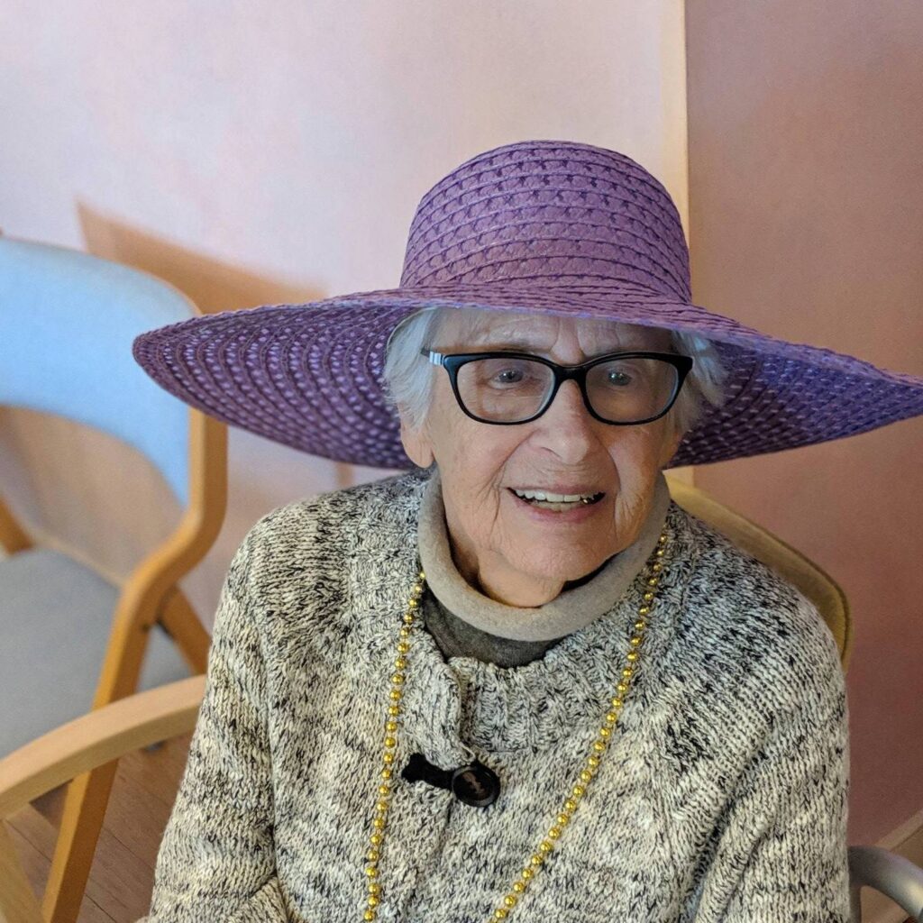 A smiling older woman in black glasses and a wide purple straw hat