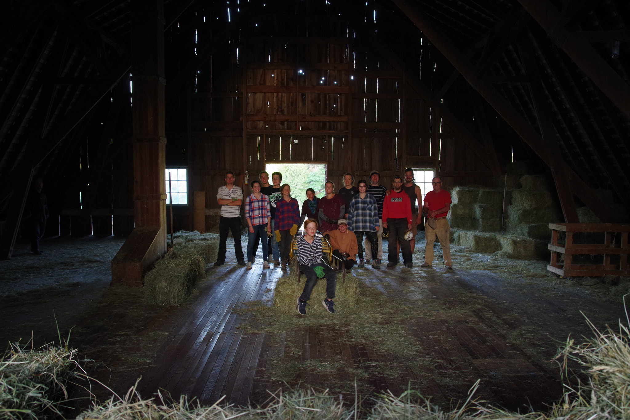 A group of people pose together inside an old barn, with light peeking through the boards.