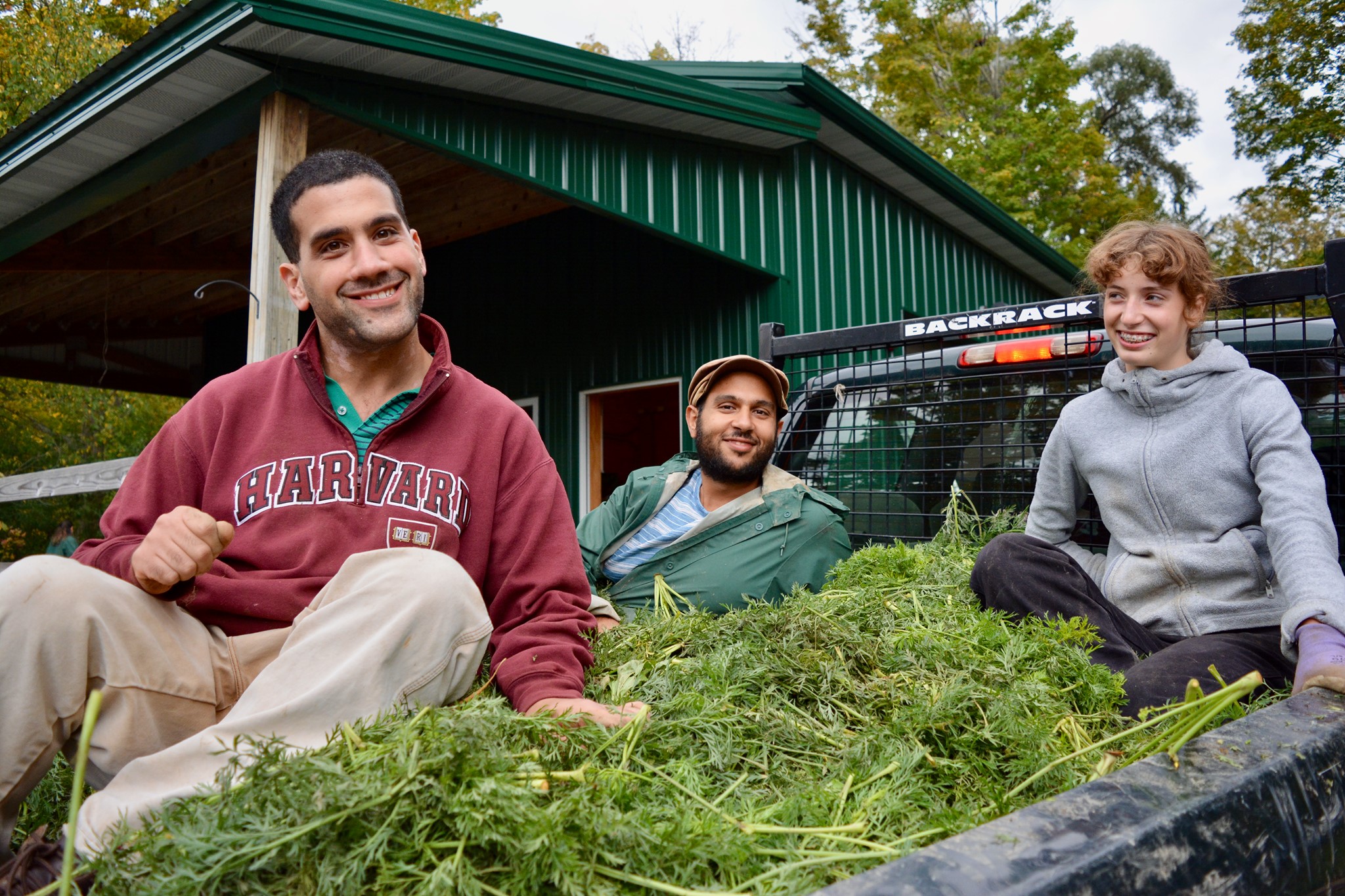 Three people sit in the bed of a truck on top of harvested greenery, smiling at the camera