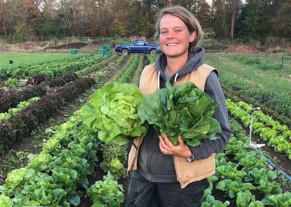 A young woman holds up two large heads of lettuce, standing in a full garden.