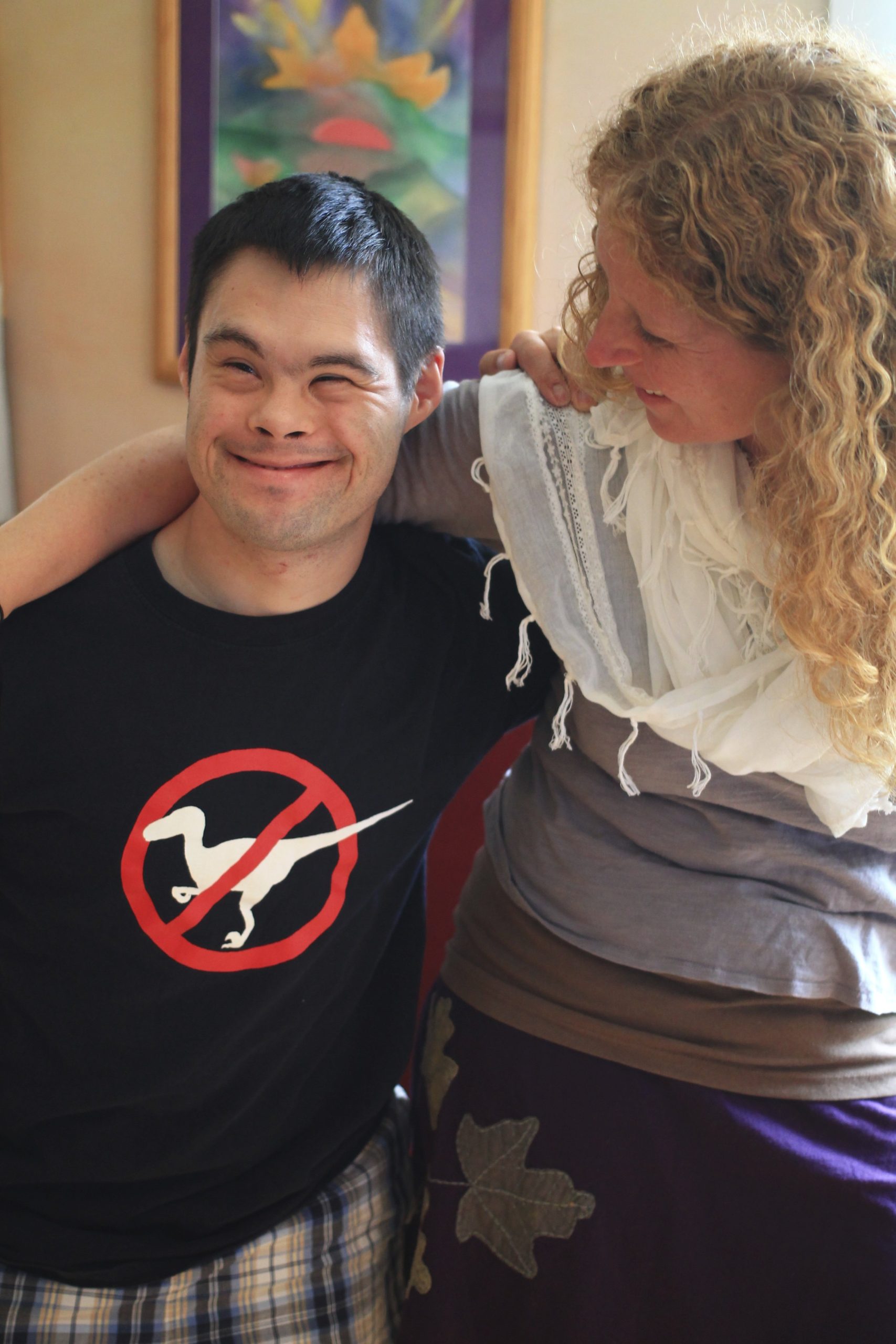 Two people hugging each other and smiling. The boy is wearing a black shirt with a silhouette of a velociraptor on it, crossed out.