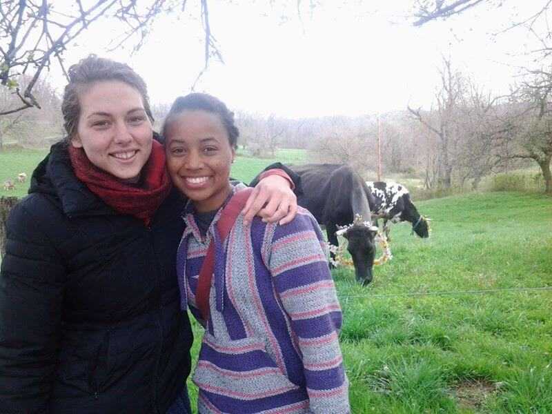 Brittany, wearing a pink and purple striped hoodie smiles with her arm around a friend who is also smiling while wearing a black coat and a red scarf. Behind them are two horses in a large field of trees and grass.