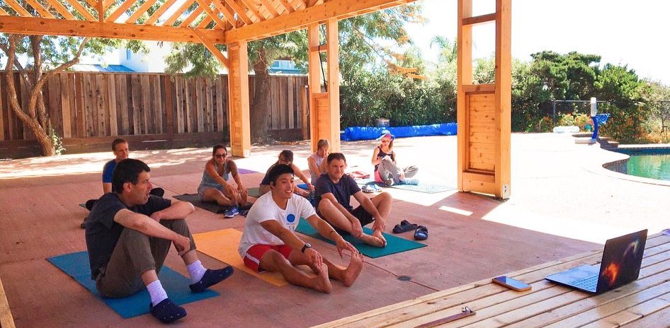 A group of people do yoga outdoors together with a video guiding them from a laptop