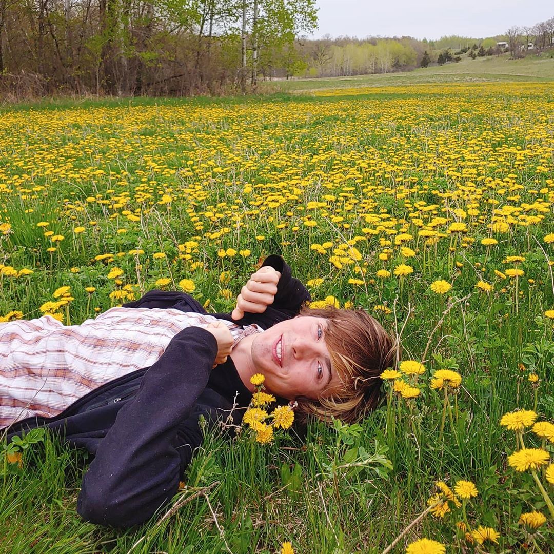 A person lies in a field of dandelions and smiles at the camera