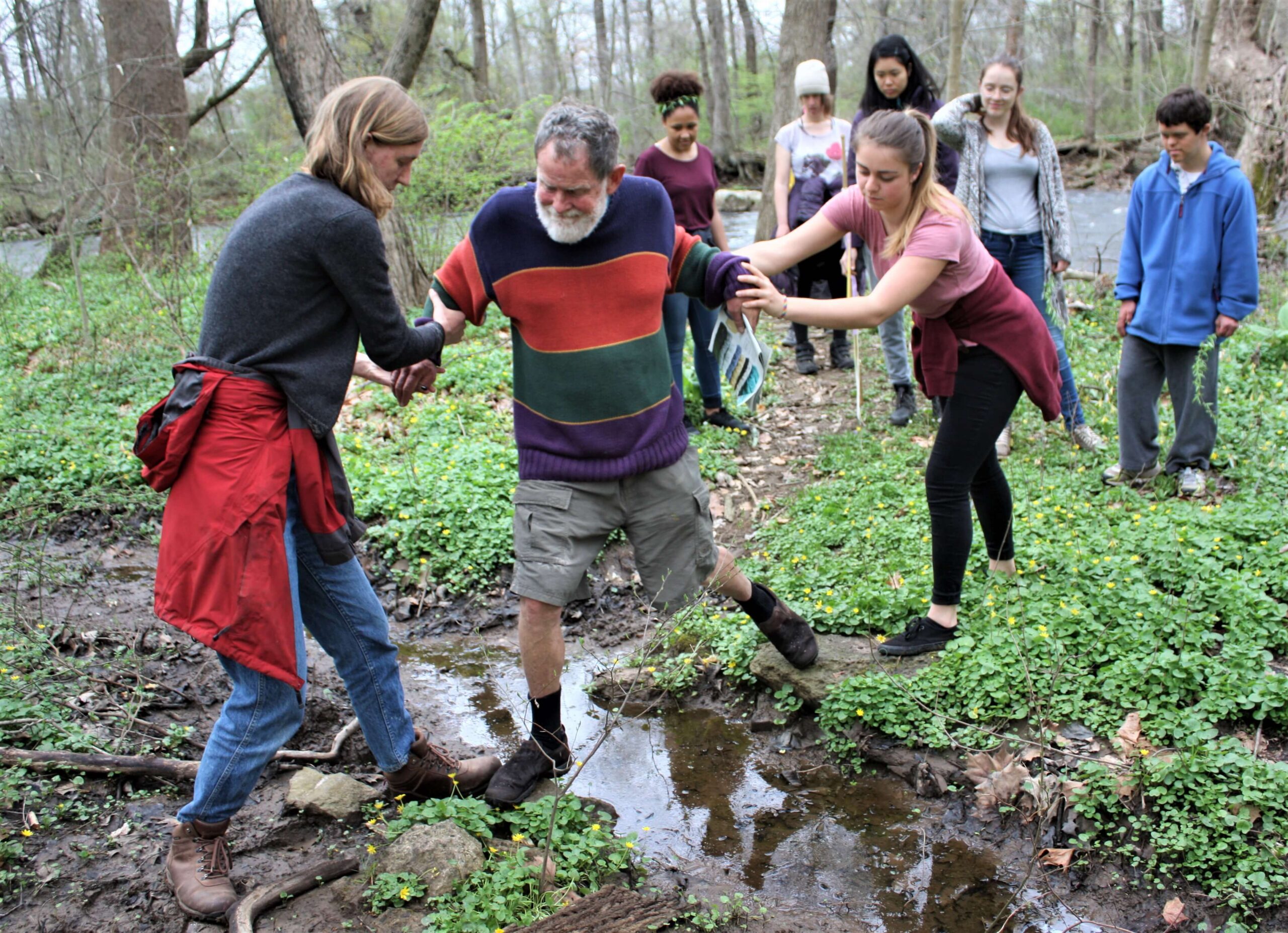A group of people walking in the woods. Two people help an older man step across a brook.