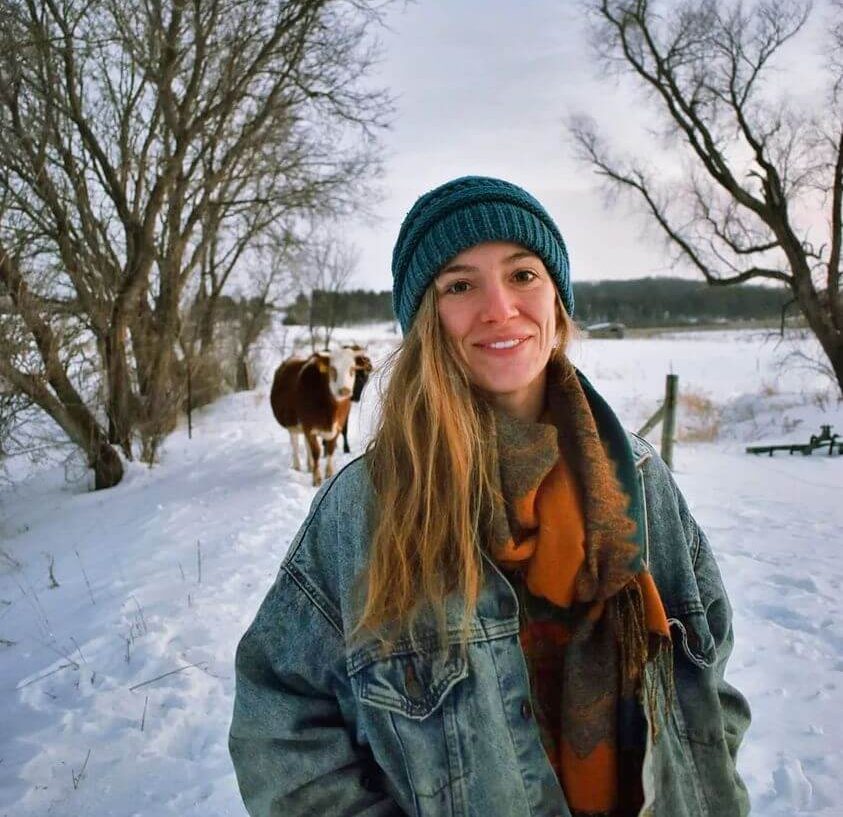 Charlotte is wearing a jean jacket, an orange and green scarf, and a blue beanie. She is standing on a snowy path with trees on either side of her, and behind her there is a brown and white cow. Charlotte is smiling.