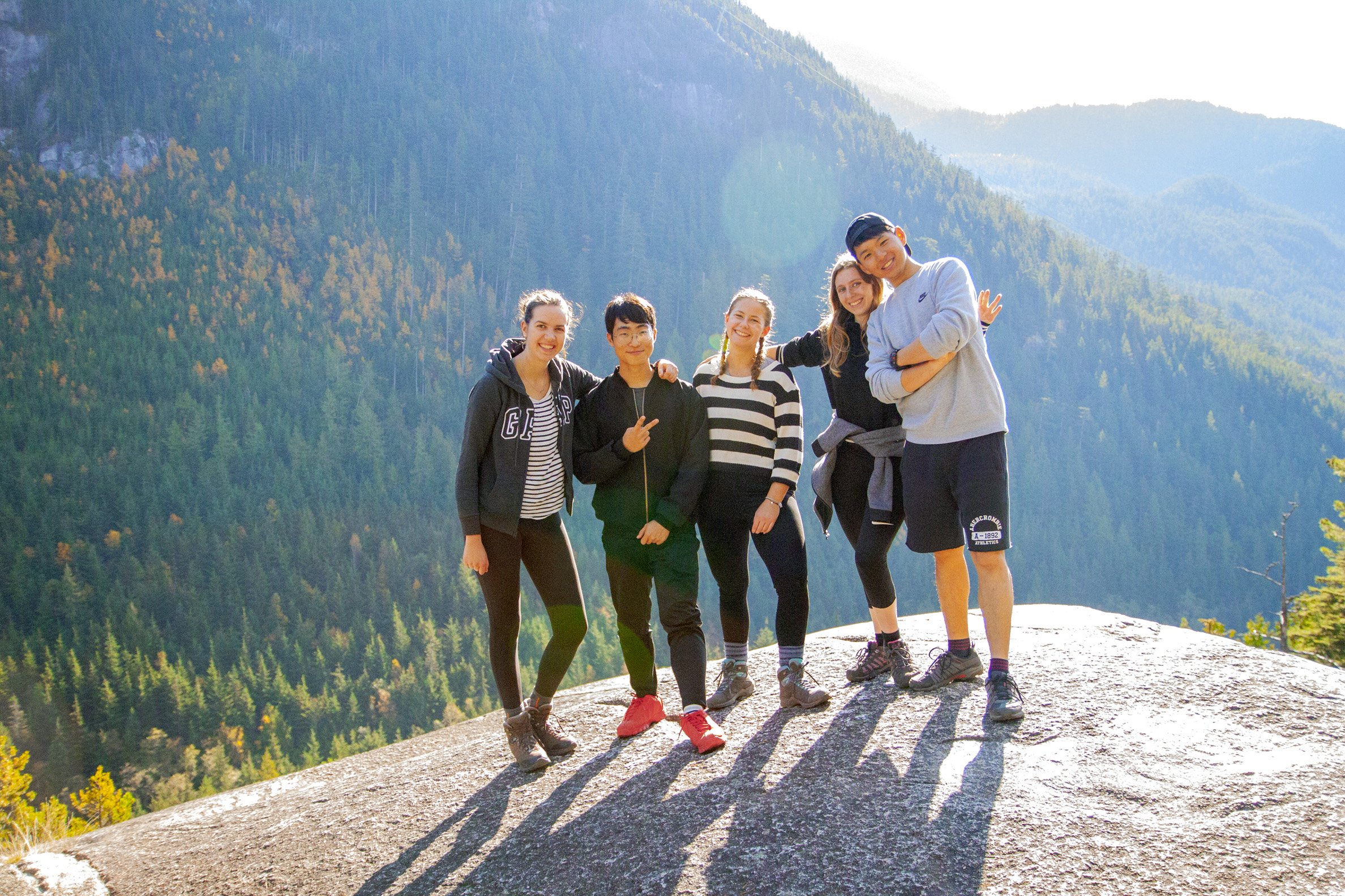 Five people in hiking clothes stand in the sunshine together on a mountain, smiling for the camera.