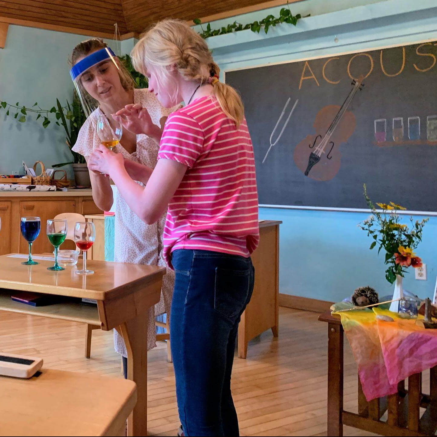 Two people demonstrate the acoustic properties of wine glasses filled with different amounts of colorful liquid in a classroom.