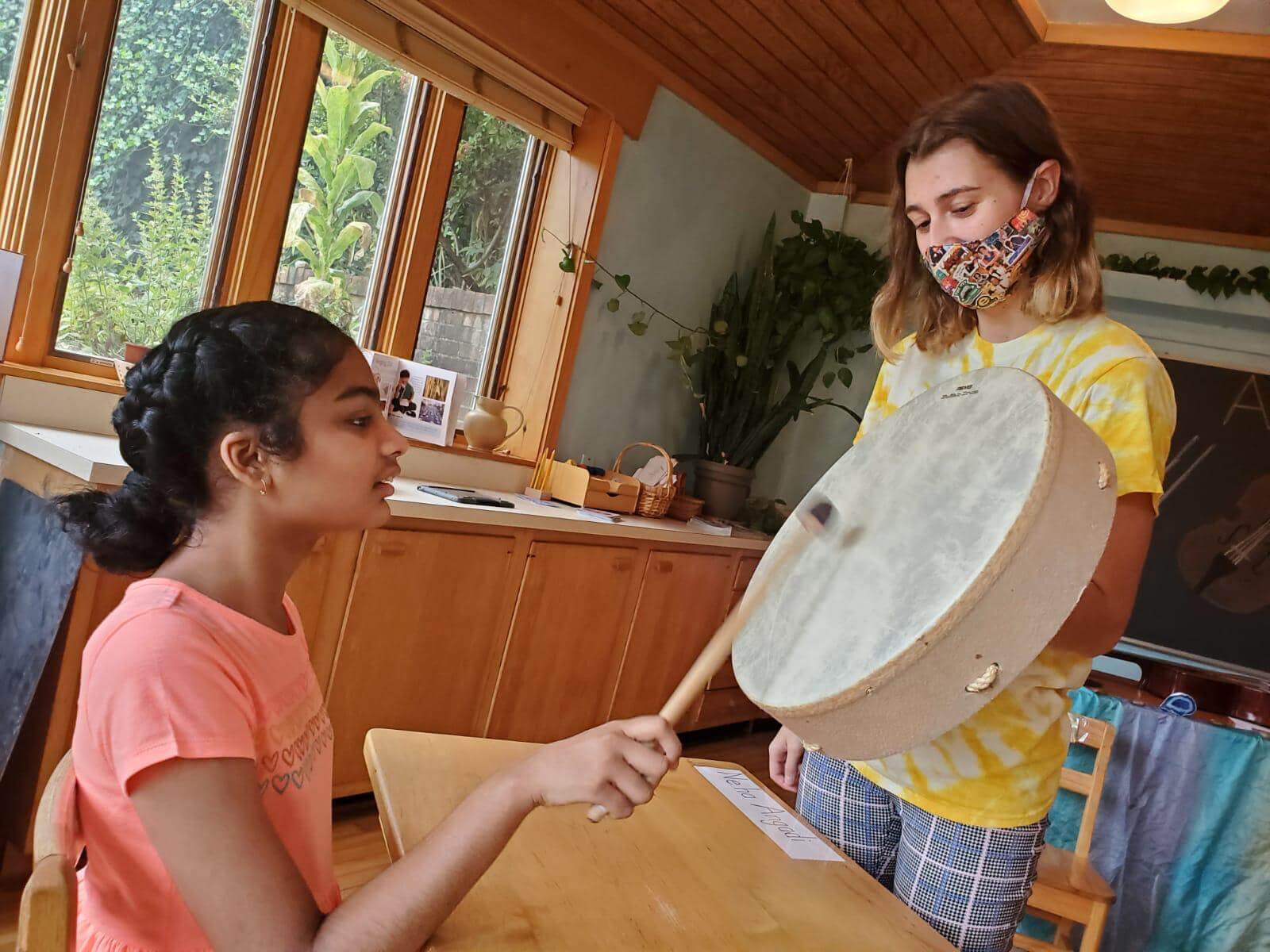 One girl holds up a drum as a seated girl taps it with a drumstick.