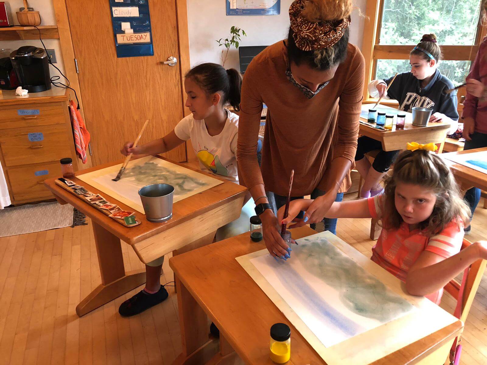 An art teacher helps students painting watercolors at their desks.