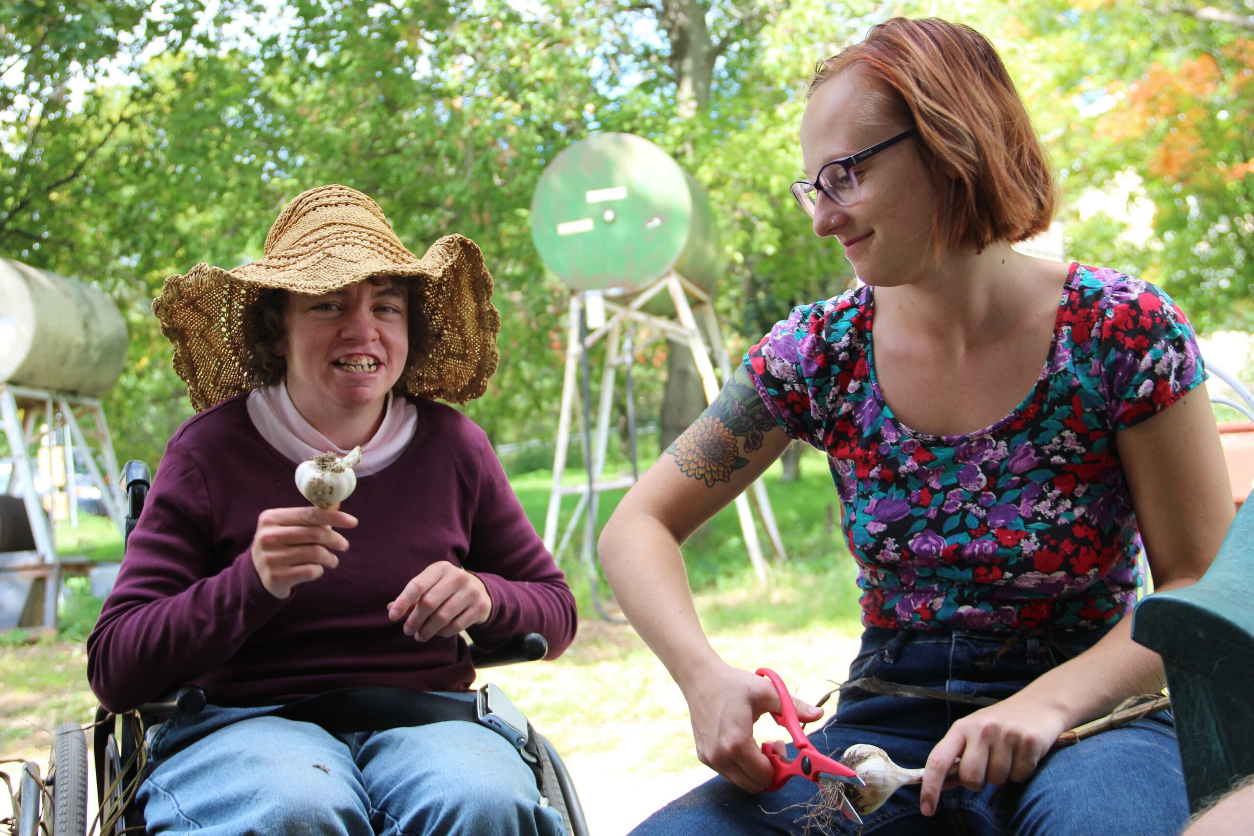 Two young women trim the roots off garlic cloves together outdoors.