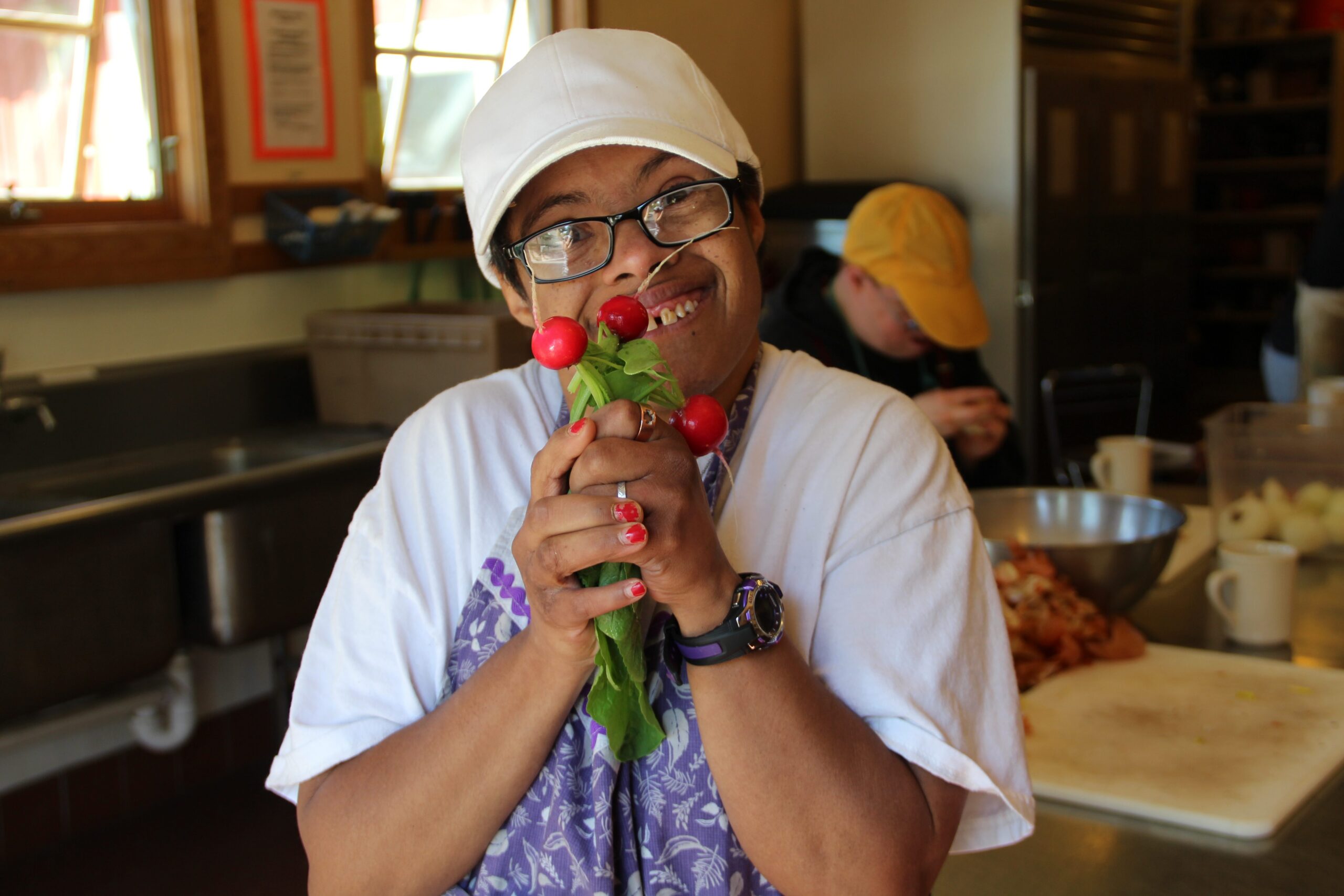 A Camphill resident in a white cap holds up a cluster of bright red radishes with delight.
