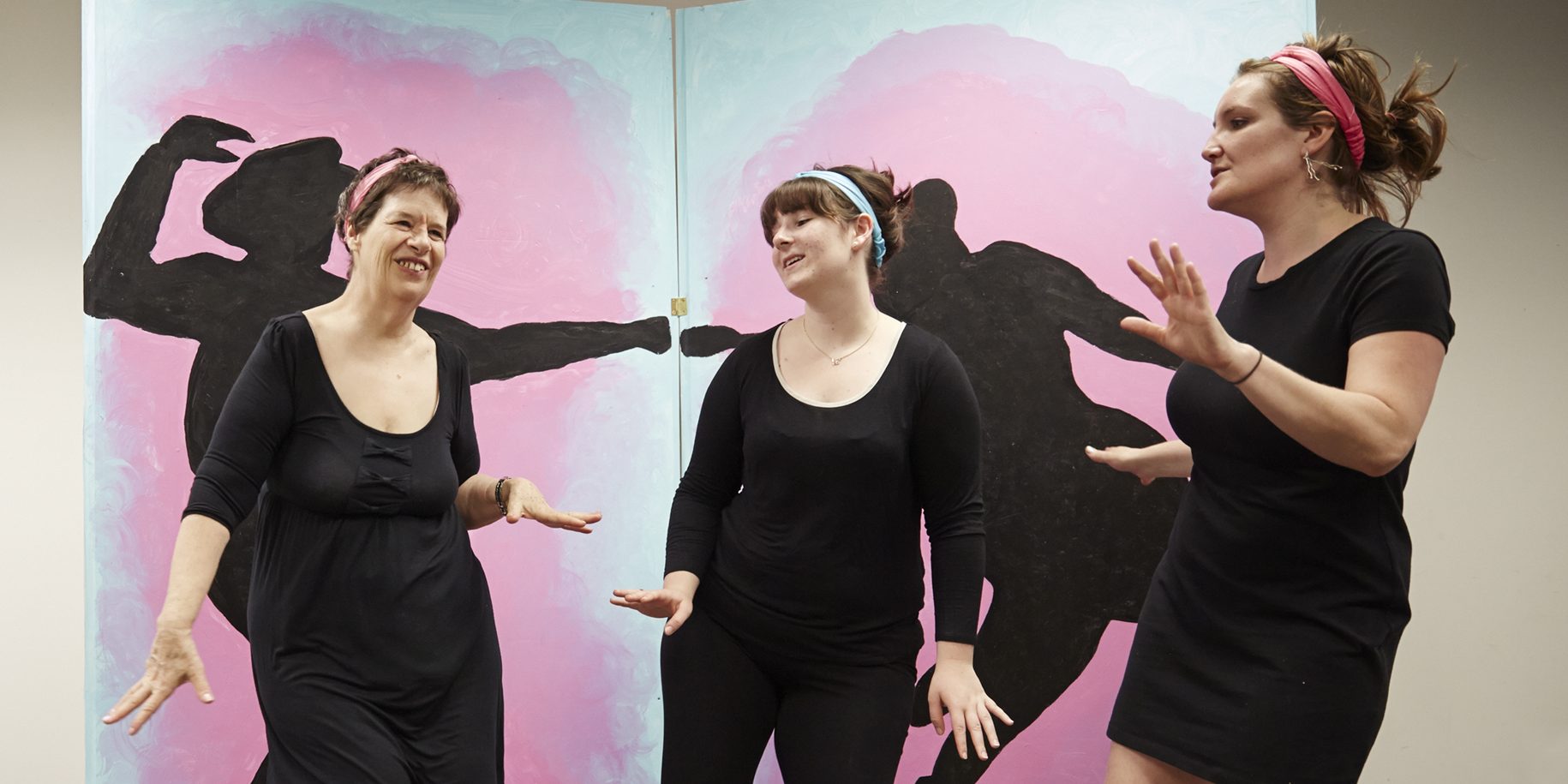 Three people in black clothes and headbands dance together in front of a mural of black silhouettes dancing.