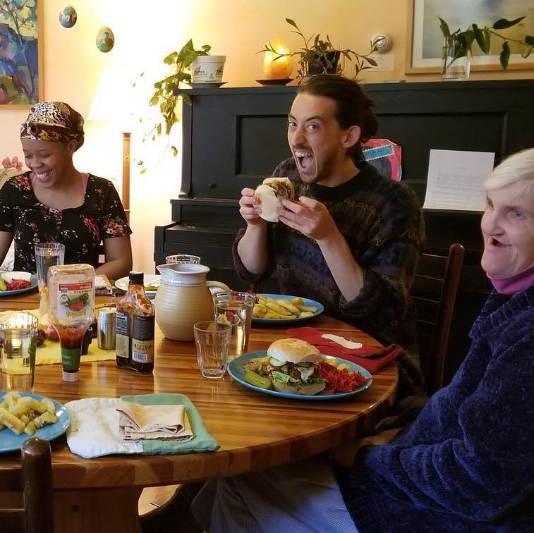 People eating dinenr together around a round wooden table. One makes a silly face at the camera as he bites into his sandwich.