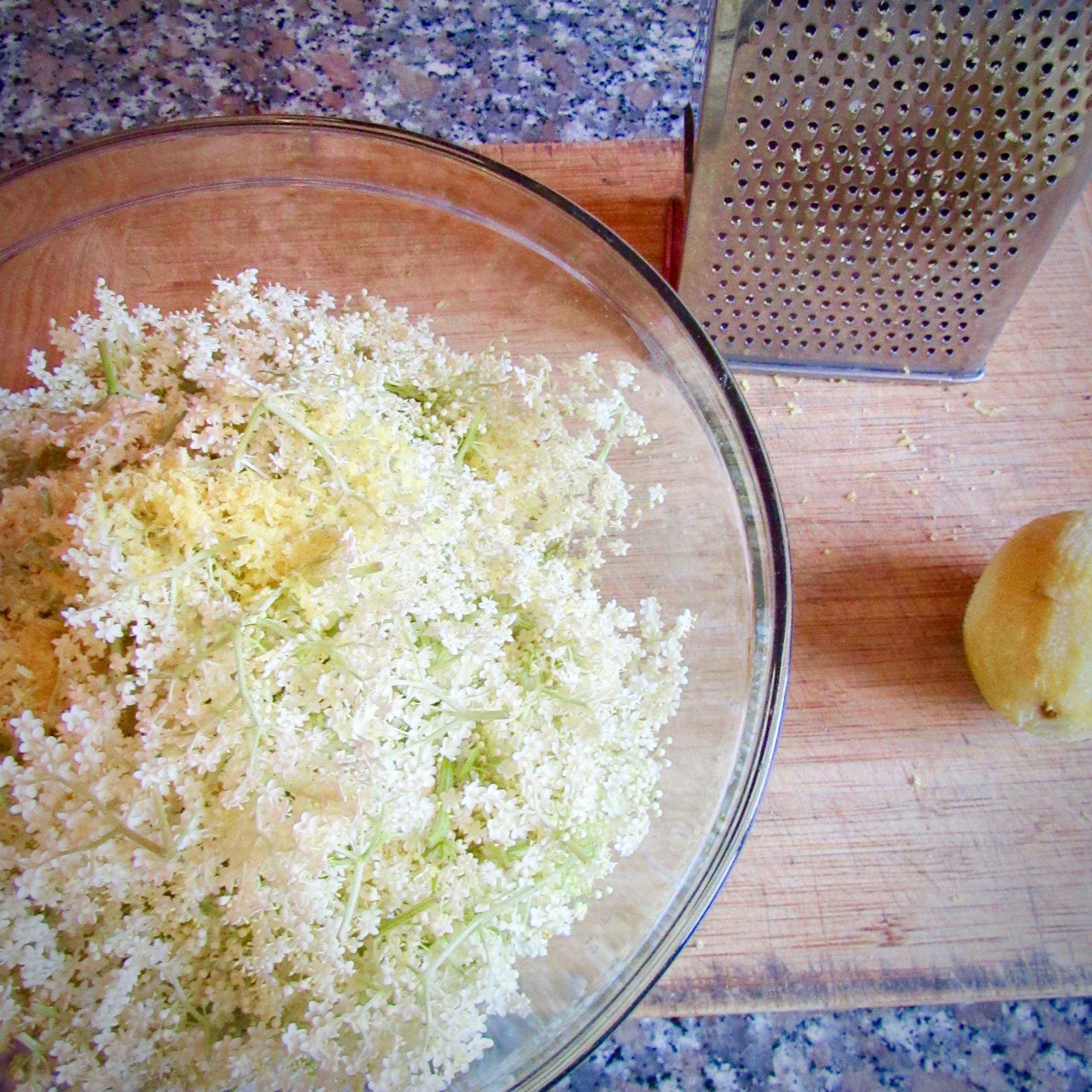 Elderflowers in a bowl on a cutting board next to a grater and a zested lemon