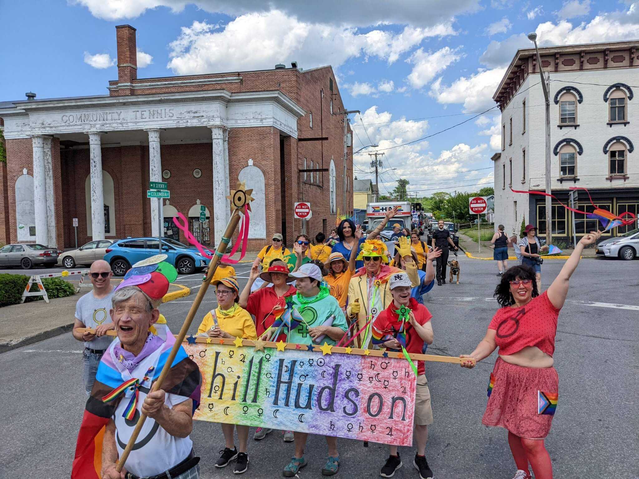 Elias is in the foreground as a crowd of people walk behind him. They are all dressed in colorful clothing and marching in a parade for Gay Pride on Warren street in Hudson New York