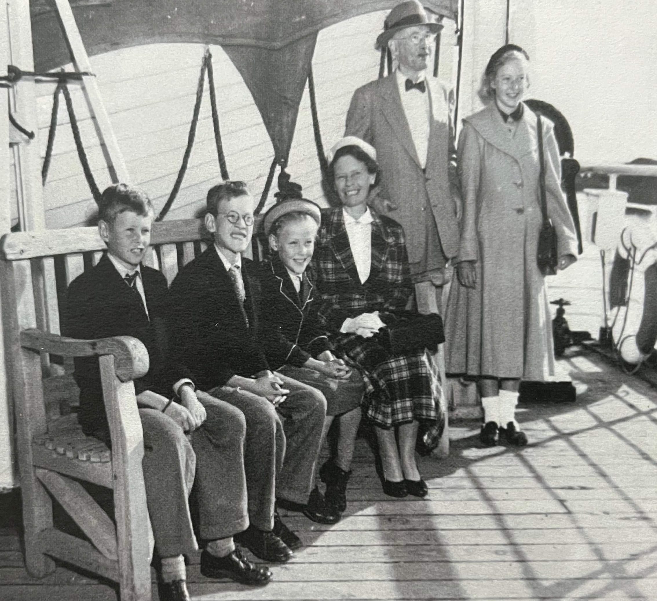 Six people dressed in formal attire pose for a press photograph on a wooden deck. The father and mother are standing on the right side and the children and nanny are sitting on a large wooden bench to the left. This image shows a young Elias and his family aboard a ship