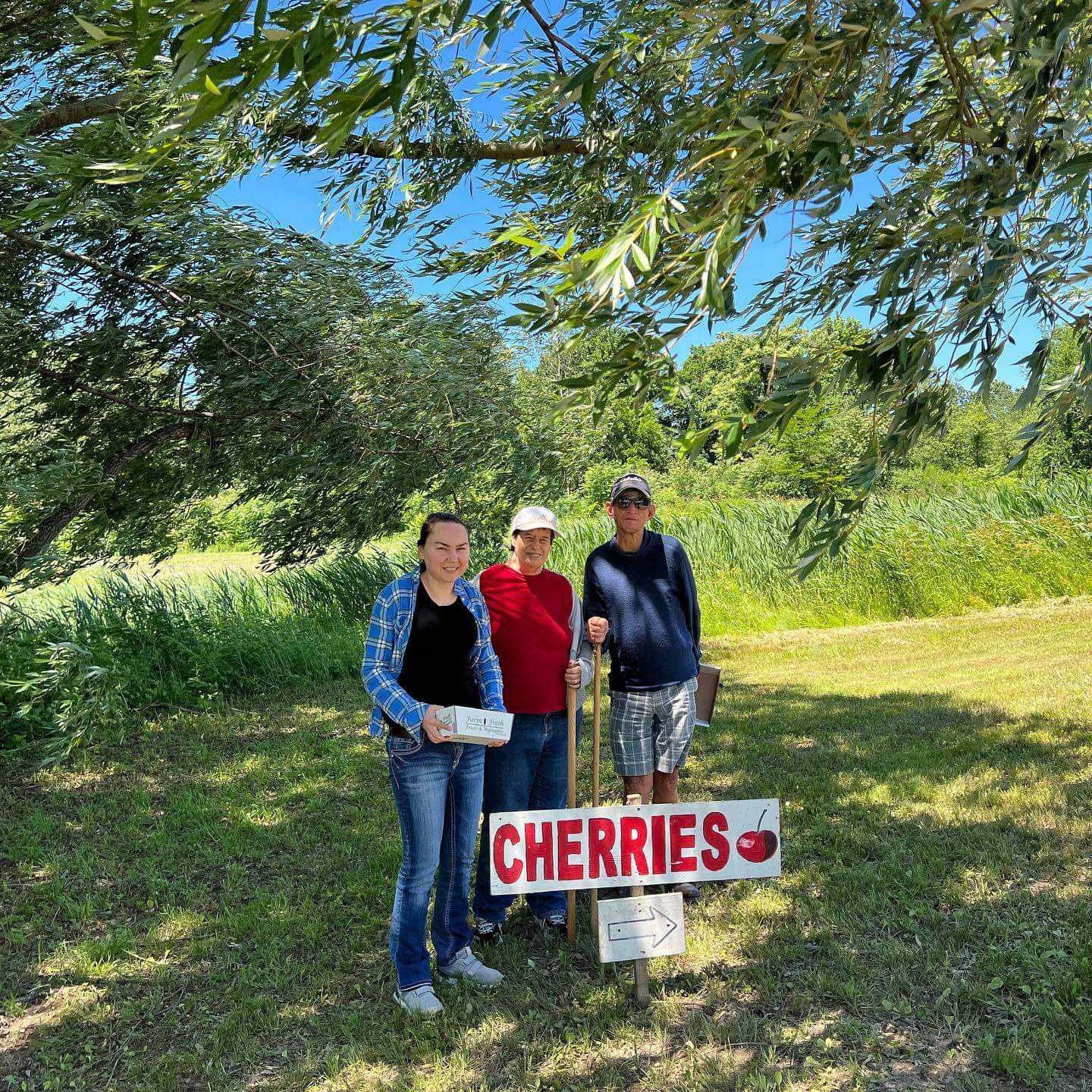 Elias stands on the right side of two of his friends. In front of them is a sign that says "cherries" and it is a beautiful sunny day