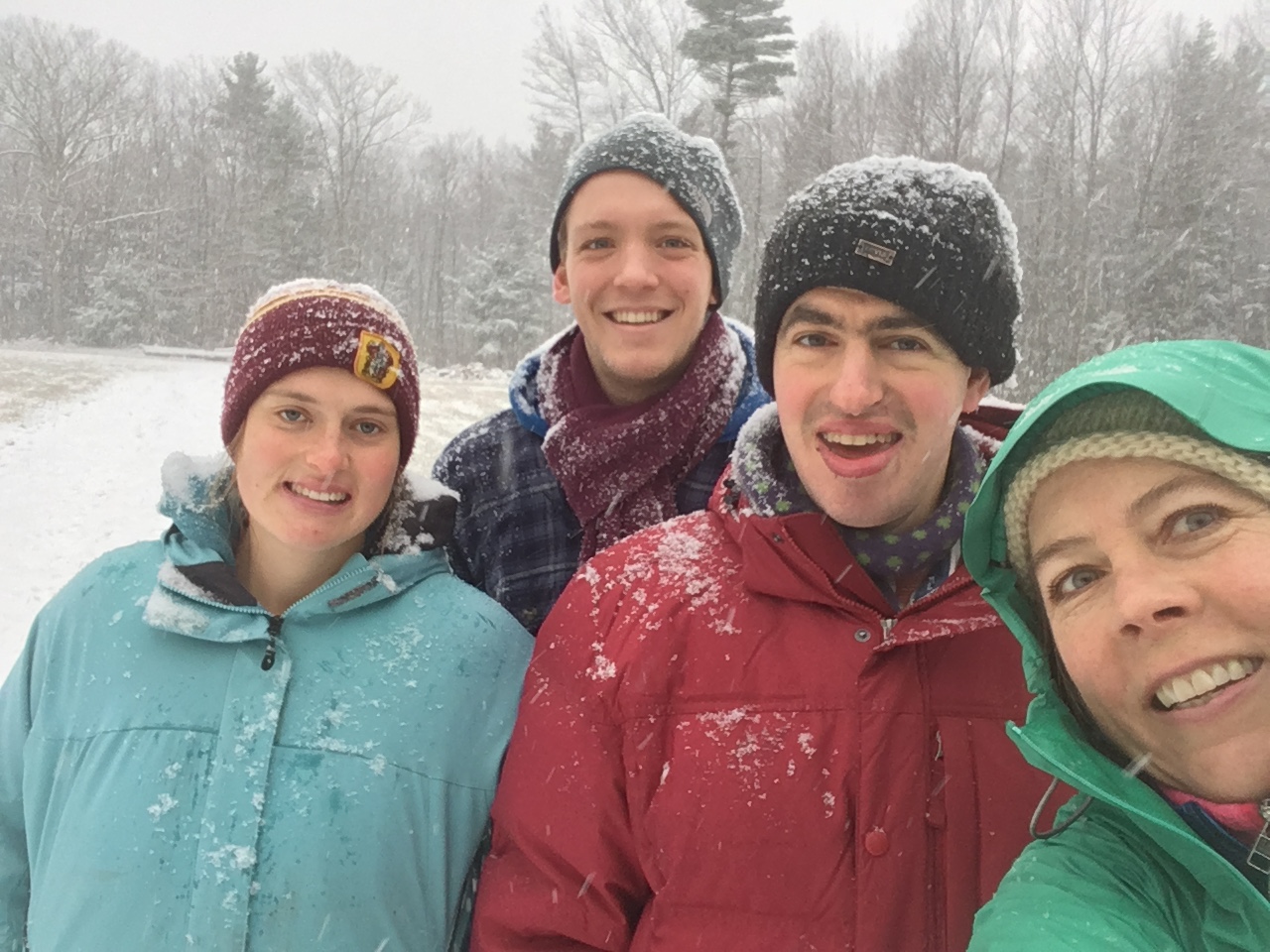 People in warm winter clothes and hats smile at the camera as they walk outside in the snow