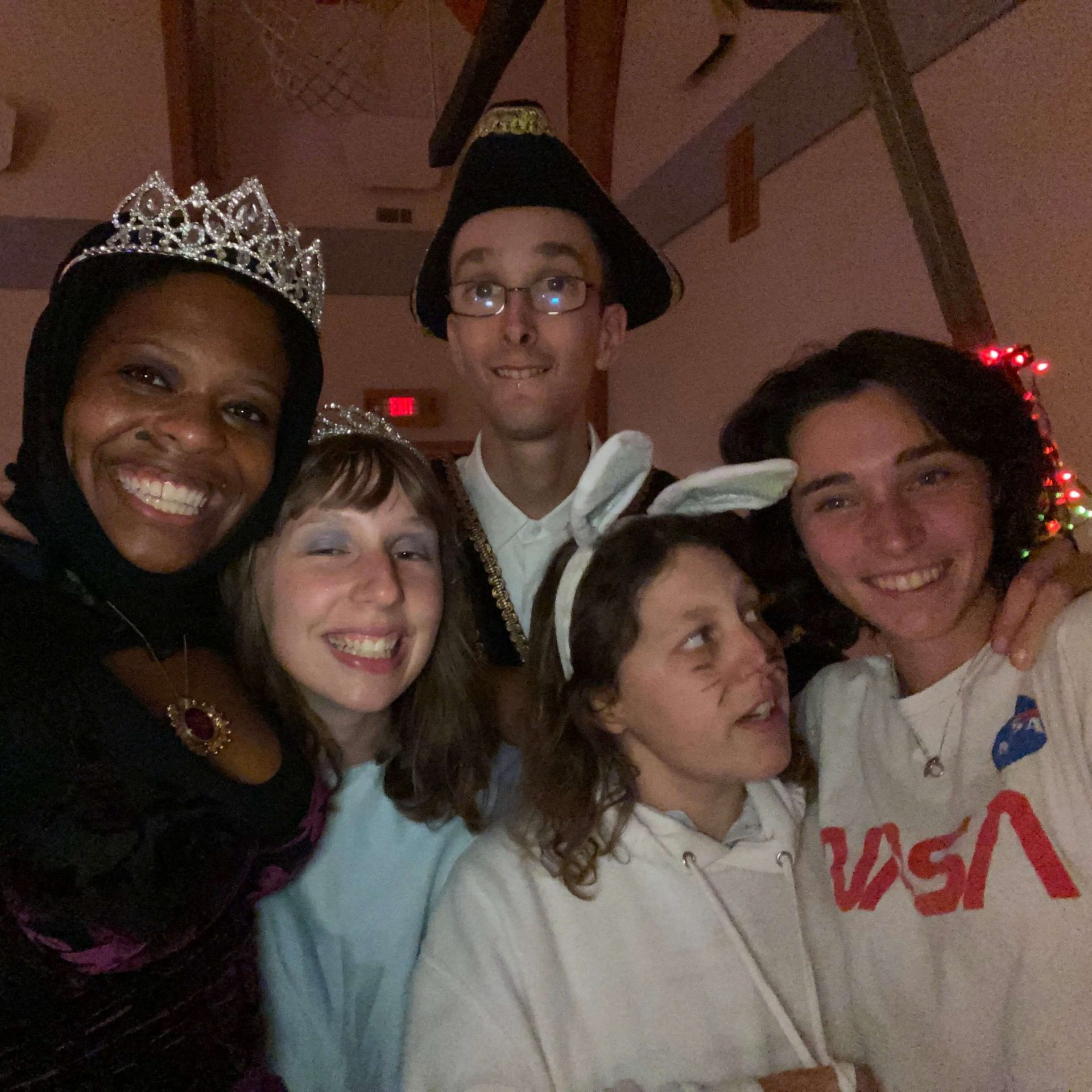 Gabi and four friends smile for the camera while dressed in costumes for Halloween. Gabi is wearing a dark purple dress and a tiara, the second friend from the left is wearing a tiara and a blue dress, the friend in the back is wearing a triangular black hat with gold stitching on it, the second friend from the right is wearing a white hoodie and white bunny ears, and the friend on the right is wearing a NASA tshirt.