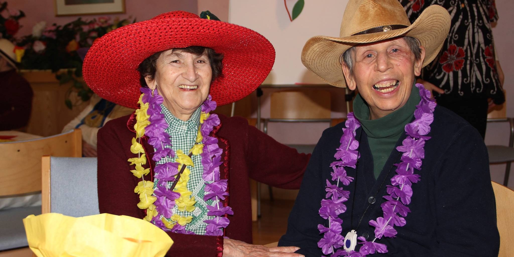 Two older adults smiling, wearing big hats and purple and yellow leis.