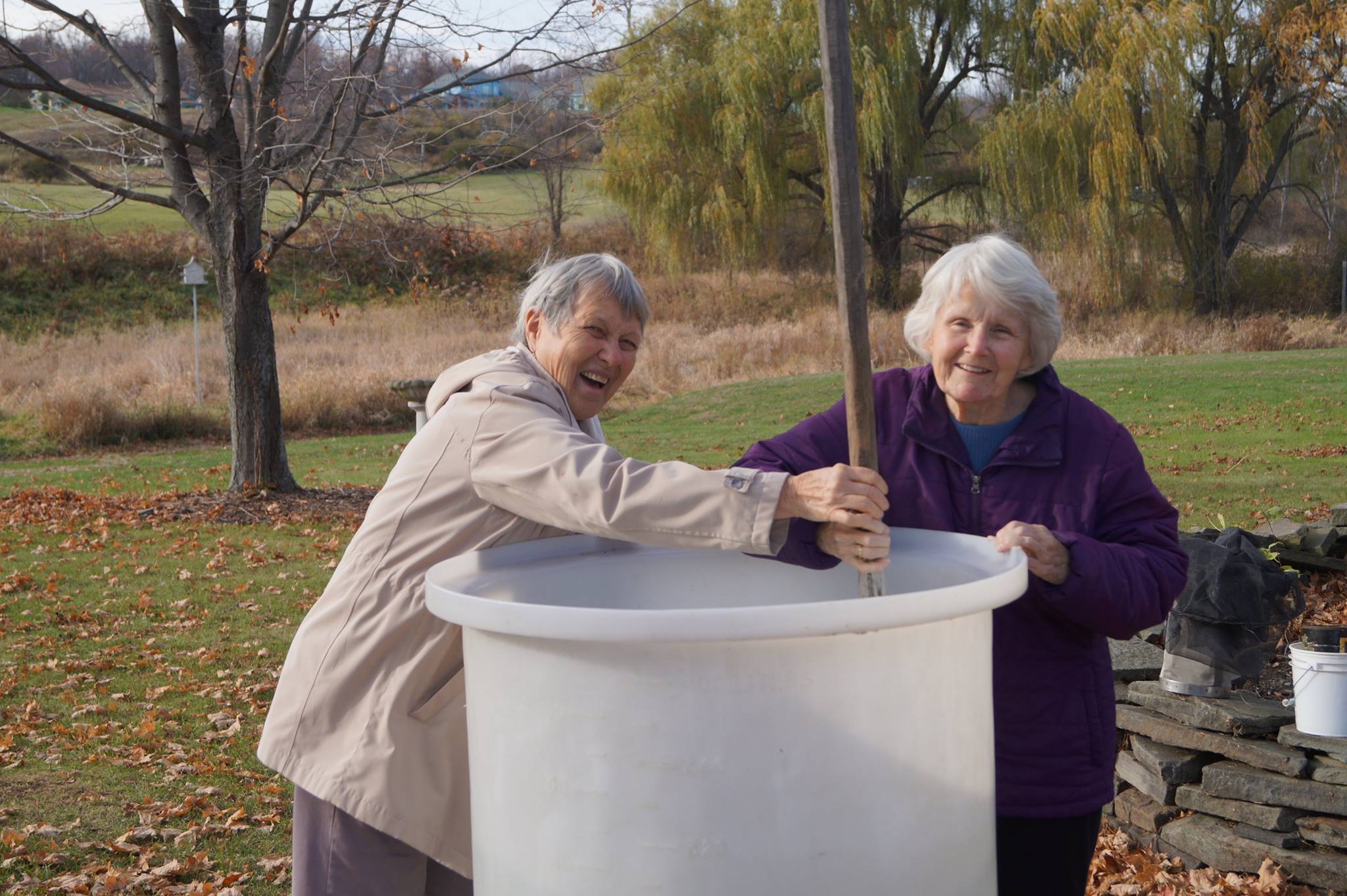 Two older women use a long stick to stir a large bucket, smiling at the camera