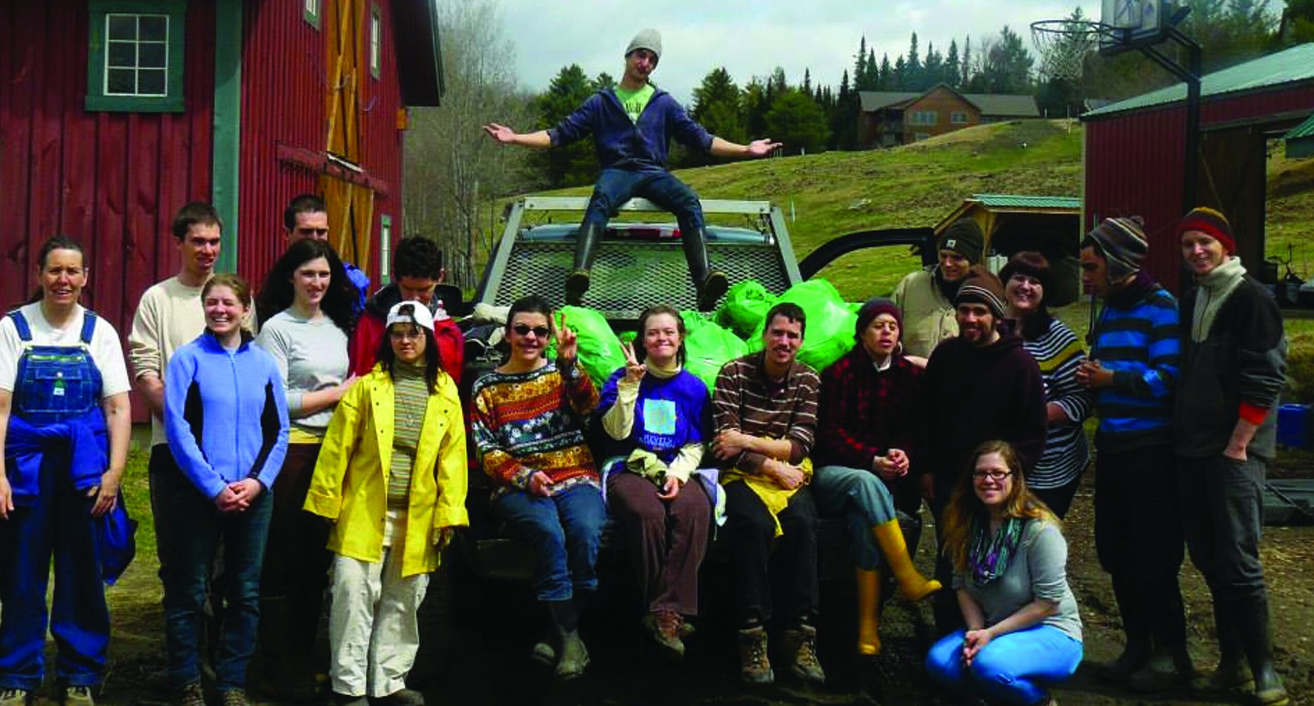 A group of people pose together on and around a pickup truck by a red barn.