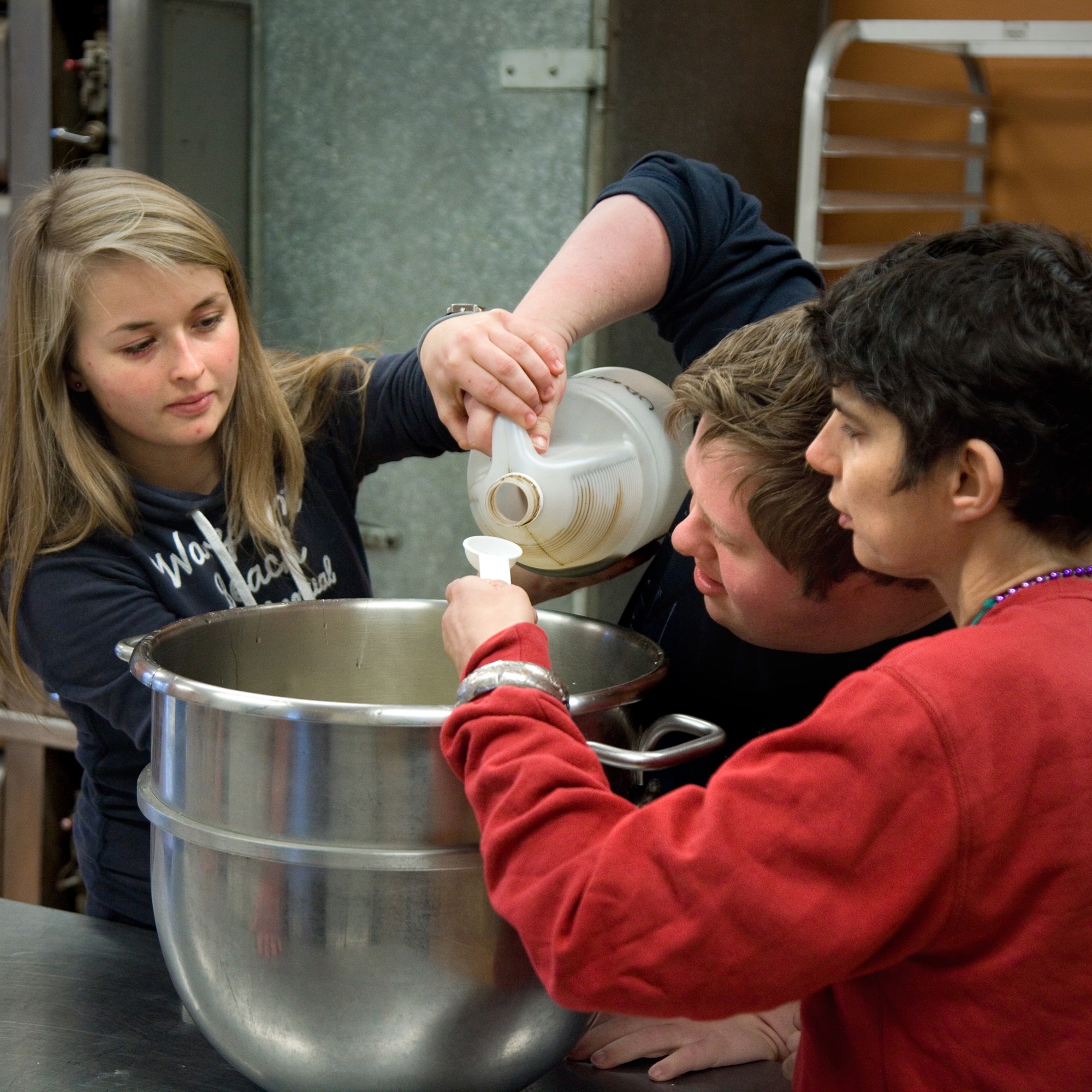 Three people in a kitchen carefully measure ingredients into a large mixing bowl