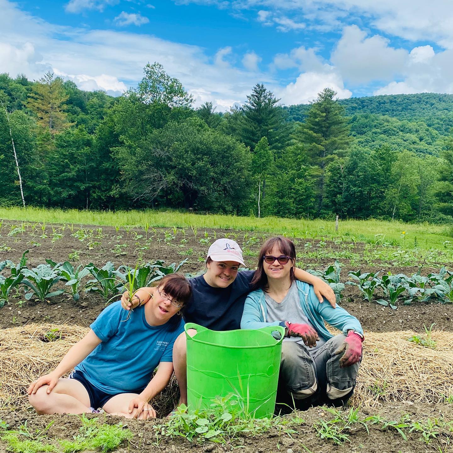 Three people sit together and pose in a garden with a large green bucket in front of them