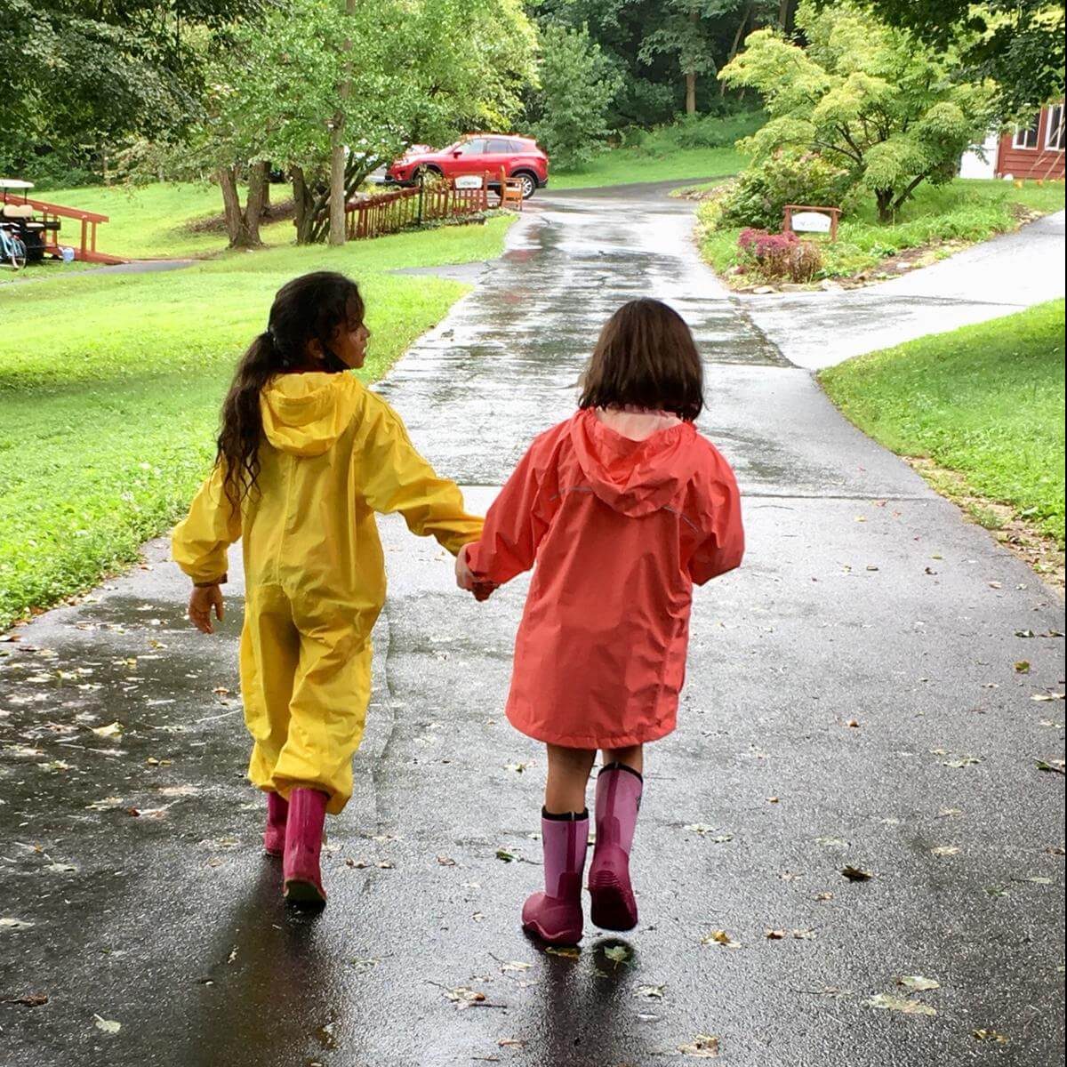 Two girls in rain gear and pink boots, holding hands as they walk down a driveway
