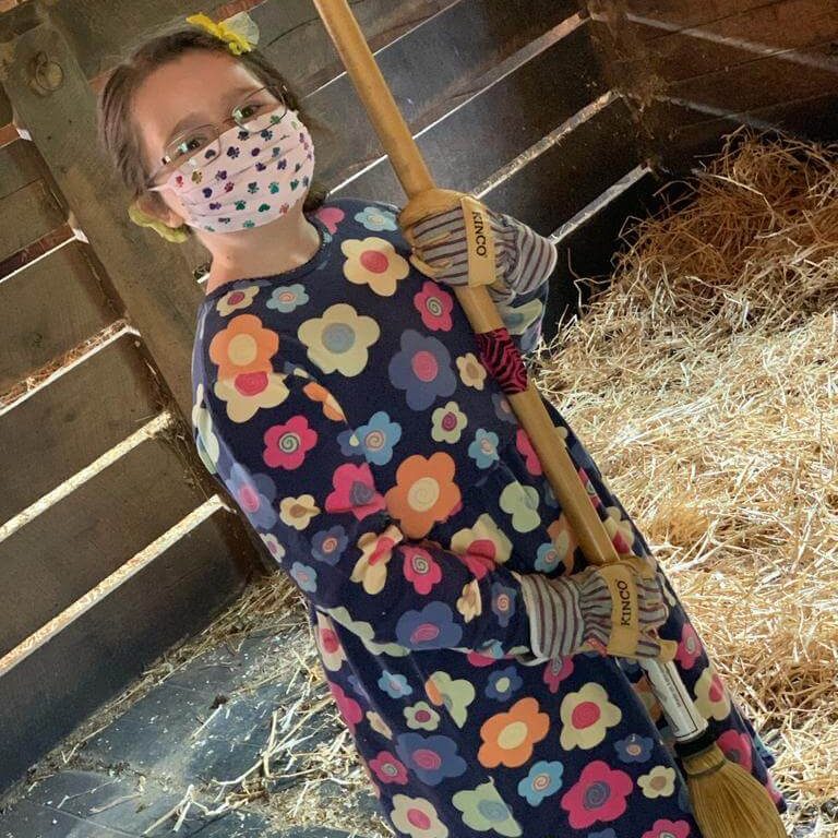 A young girl wearing a face mask and gardening gloves, holding a broom in a barn.