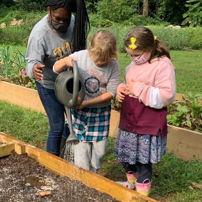 Two little girls water a raised bed with a large watering can as an adult supervises.