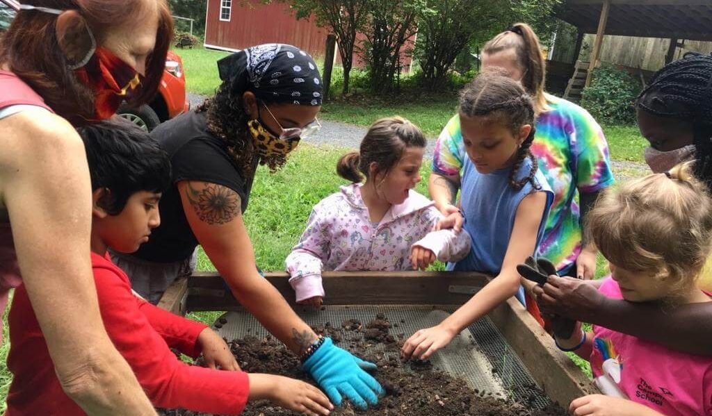 People of all ages, some with face masks and some without, filtering soil through a wire screen.