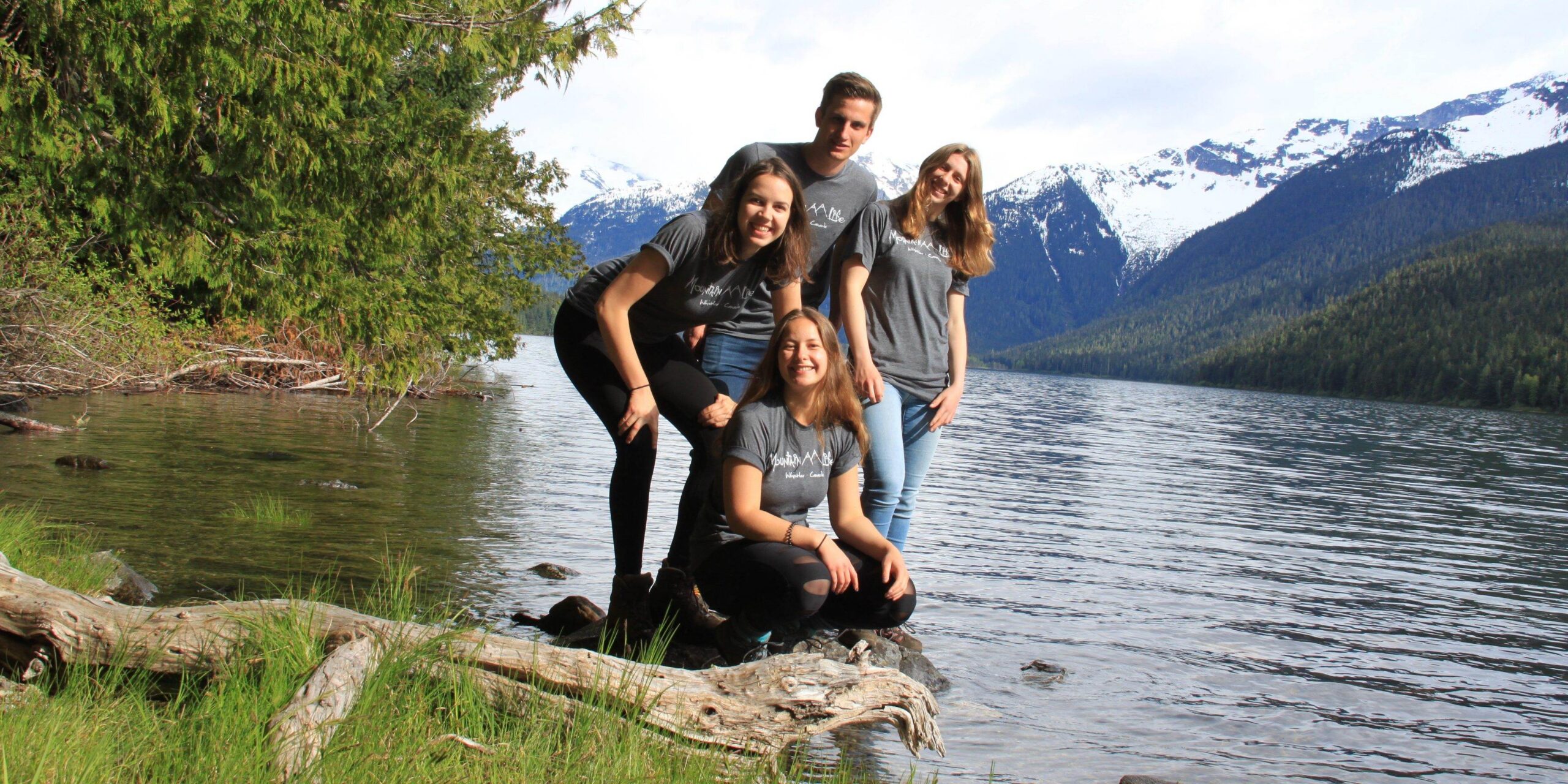 Four people in matching gray shirts pose together on a small rock by the bank of a wide river, with mountains in the background