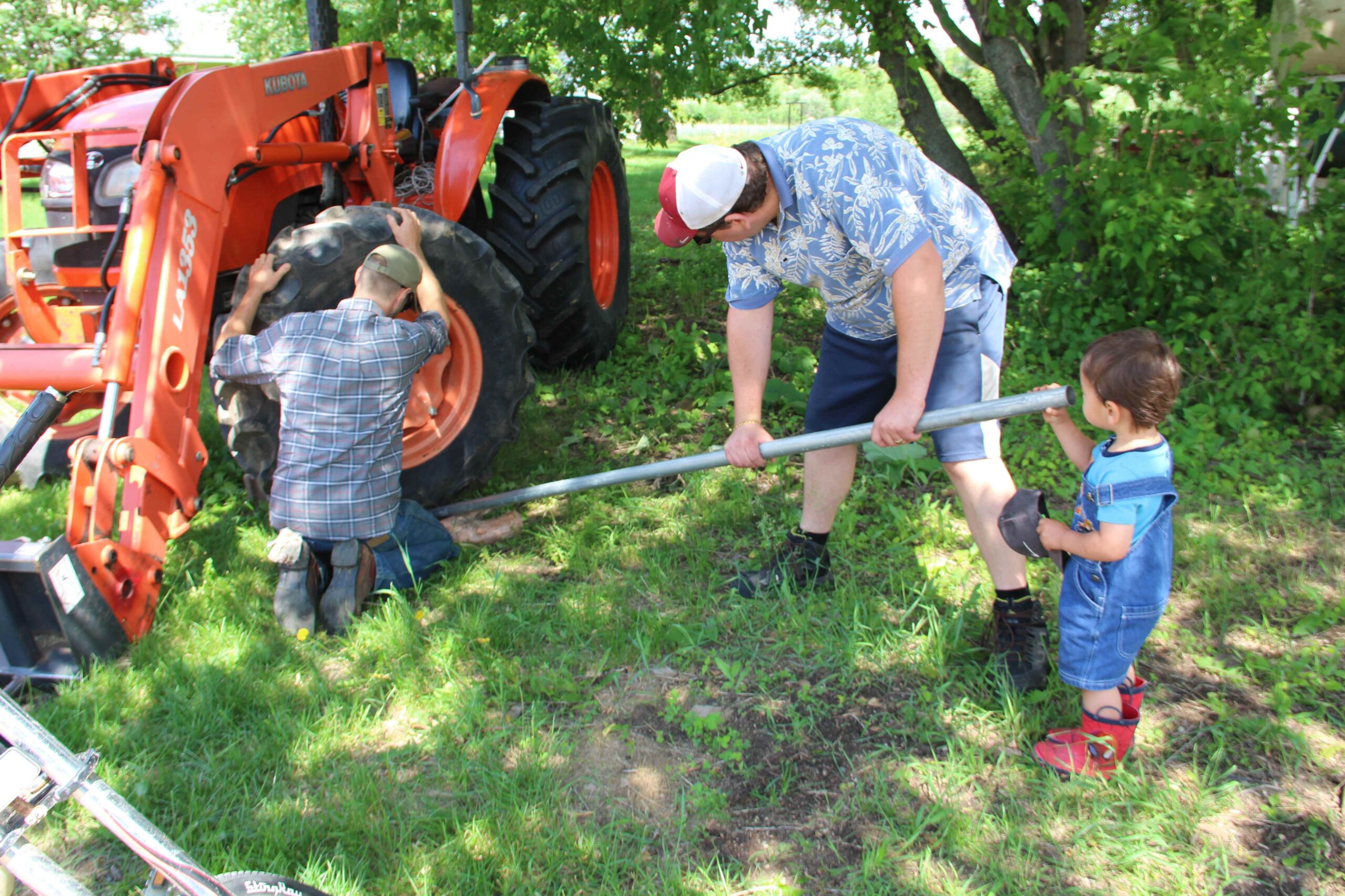 Two men work on a tractor as a toddler in red boots inspects a metal pipe.