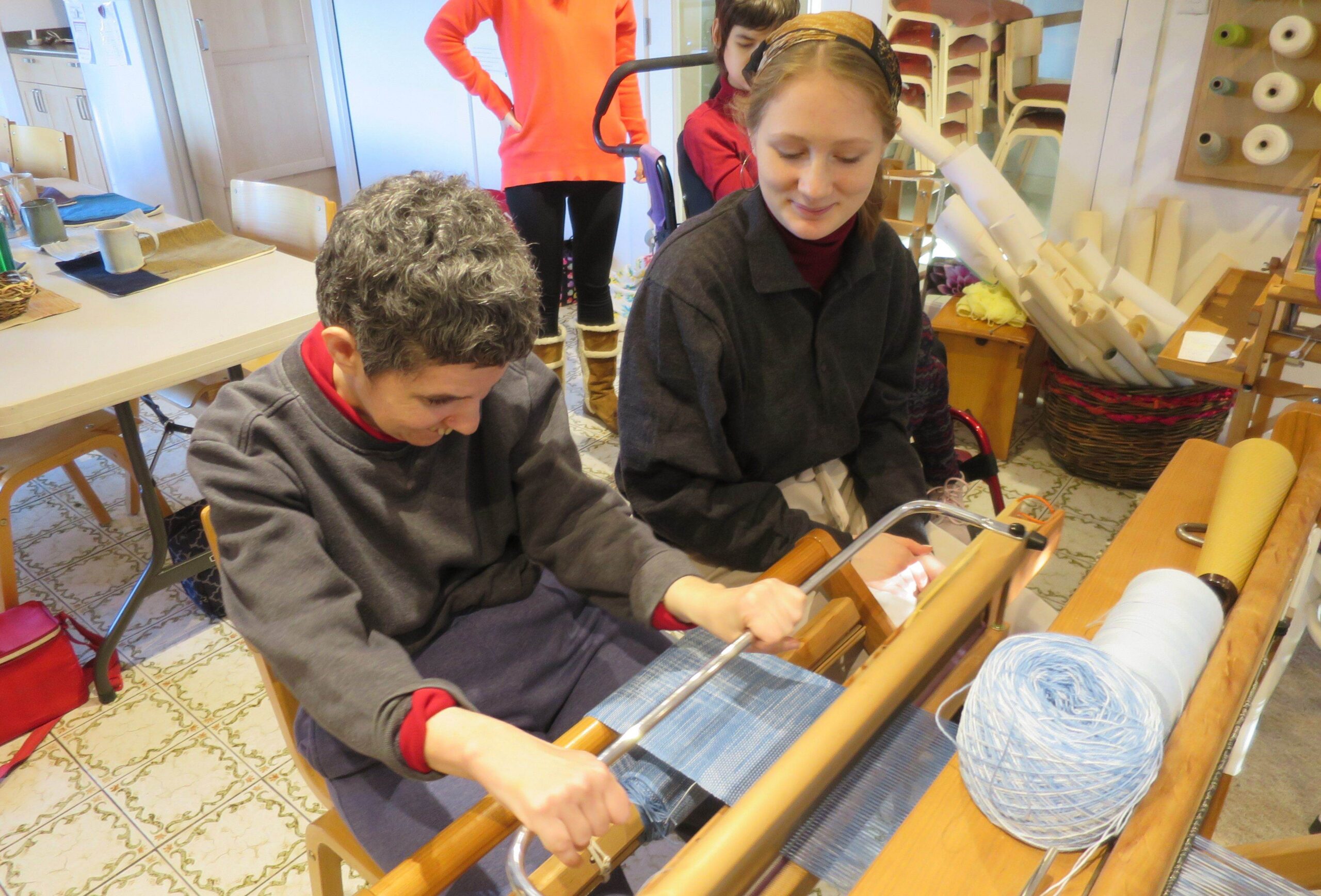 One person weaves light blue fabric on a floor loom as another looks on