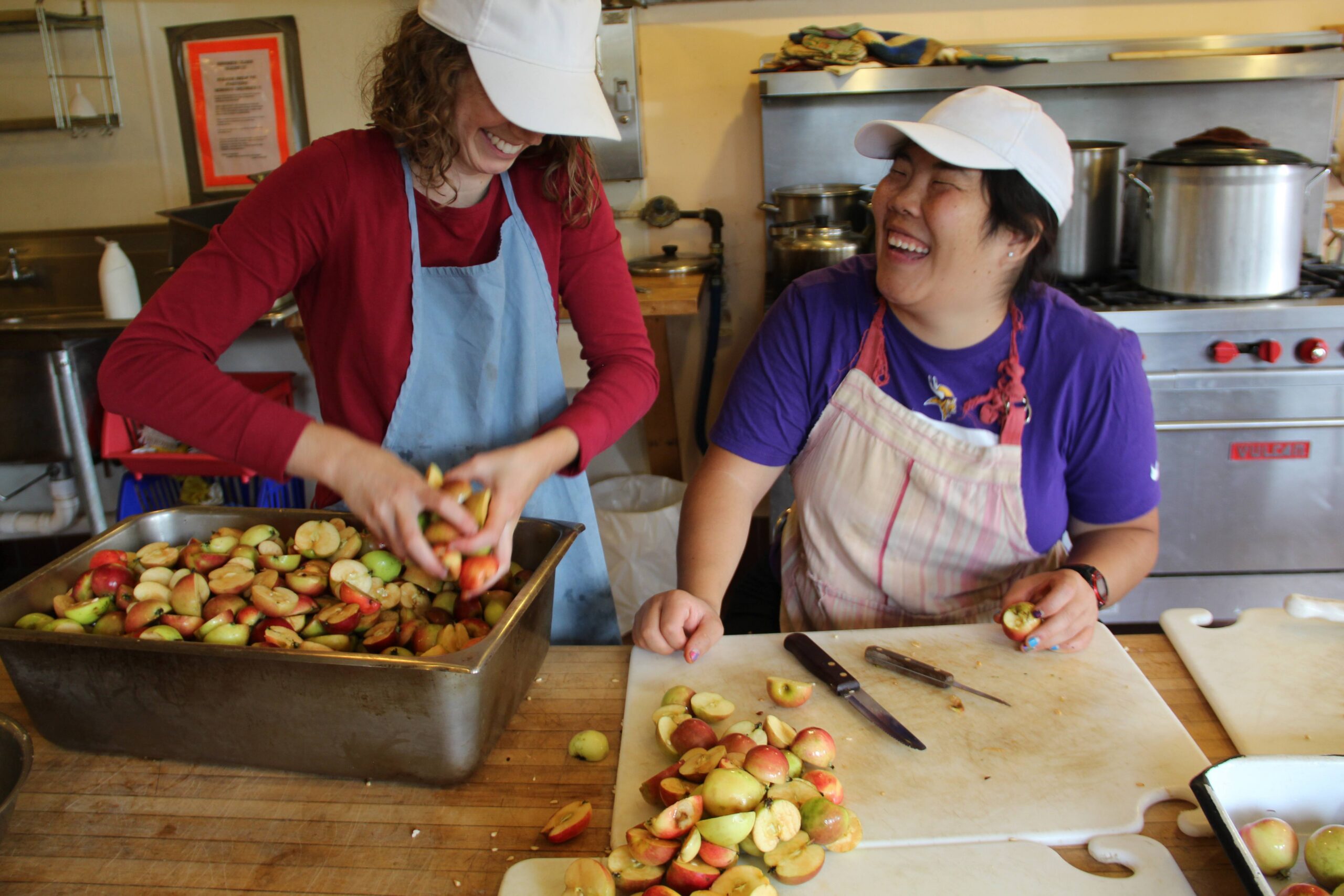 Two women laughing together as they cut up fruit in a kitchen.