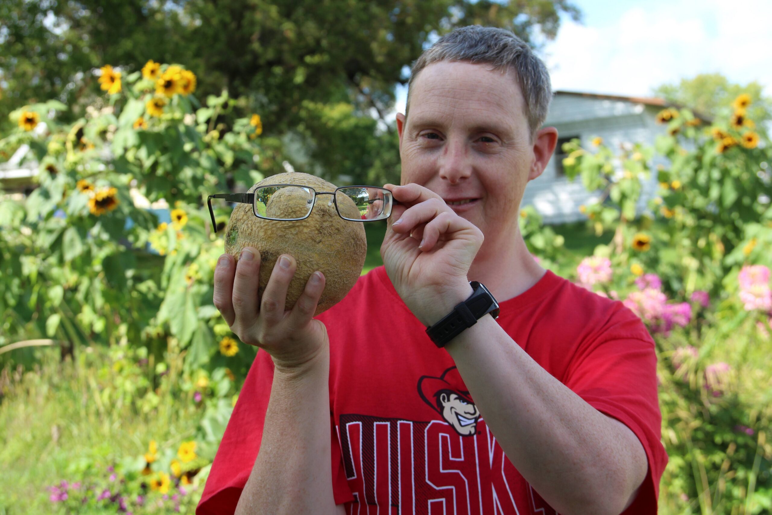 A man holds up his glasses in front of a small cantaloupe, as if the cantaloupe is wearing them.