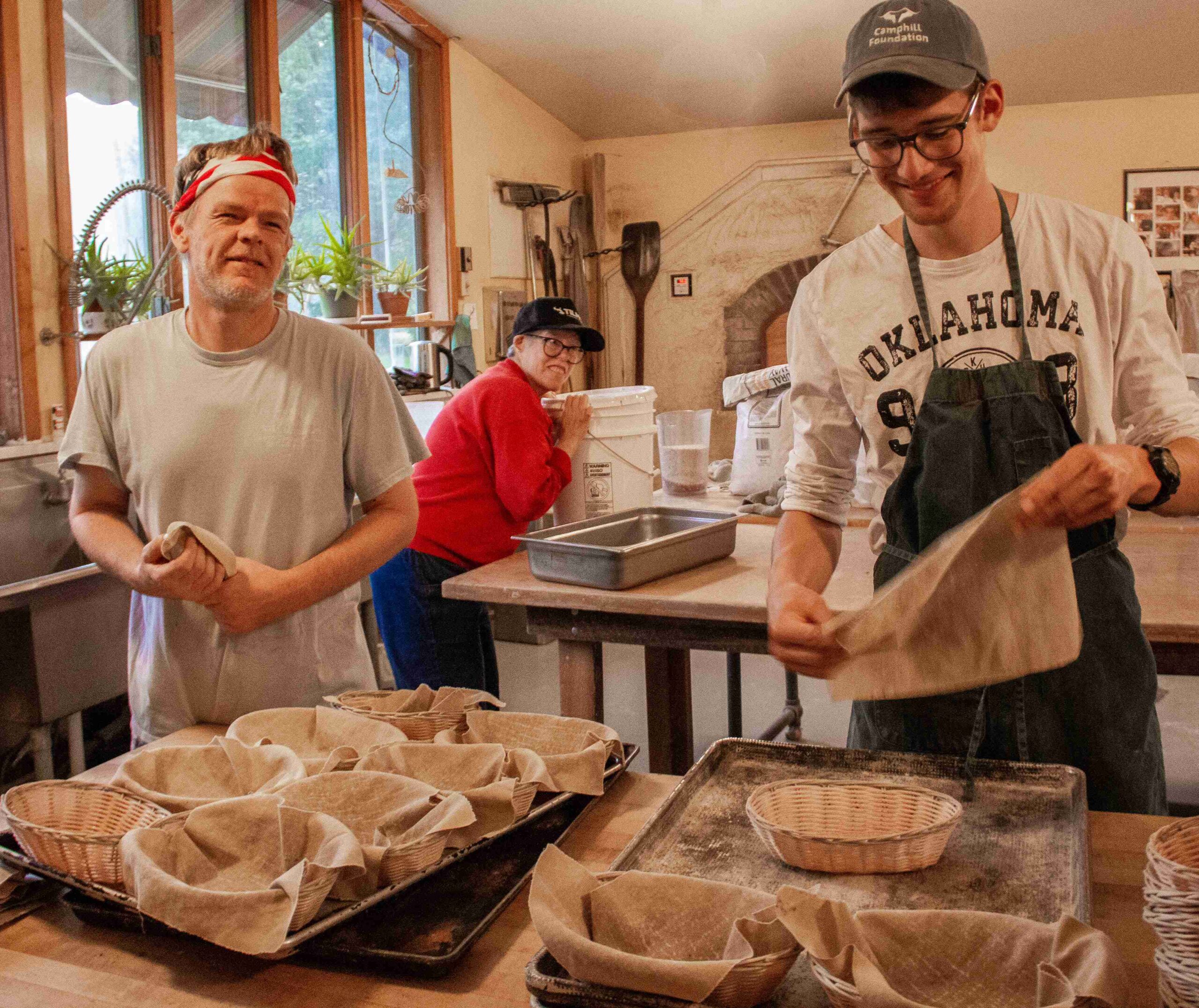 Two men line baskets with small cloth napkins as another resident lifts a white bucket in the background.