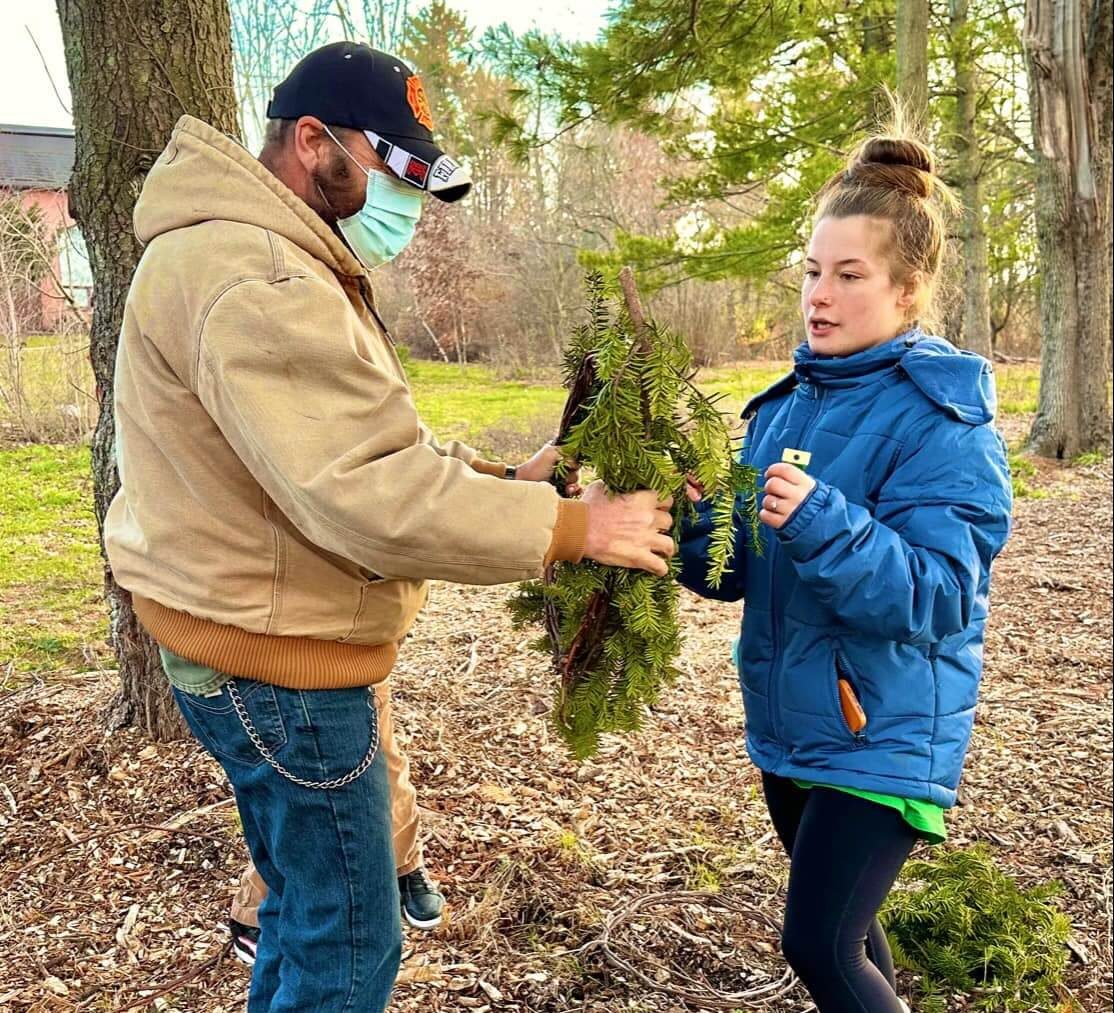 Two people stand in a field of trees and fallen leaves, working together on a wreath. The person on the right is wearing brown fuzzy boots, black pants, and a blue coat with a green shirt poking out from under it, and their light brown hair is tied in a bun on top of their head. The person on the left is wearing brown boots, jeans with a chain on the hip, a light brown coat, a green face mask, and a black and orange hat.